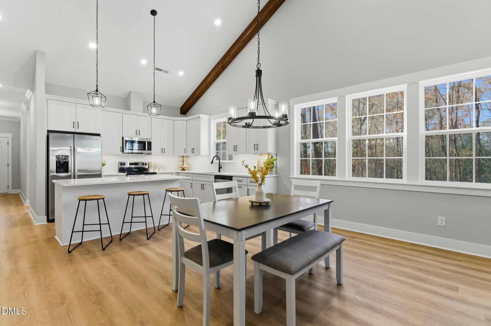 A modern kitchen and dining area with white cabinets, stainless steel appliances, and a dining table with chairs and a bench. Large windows let in natural light, and pendant lights hang from the ceiling.
