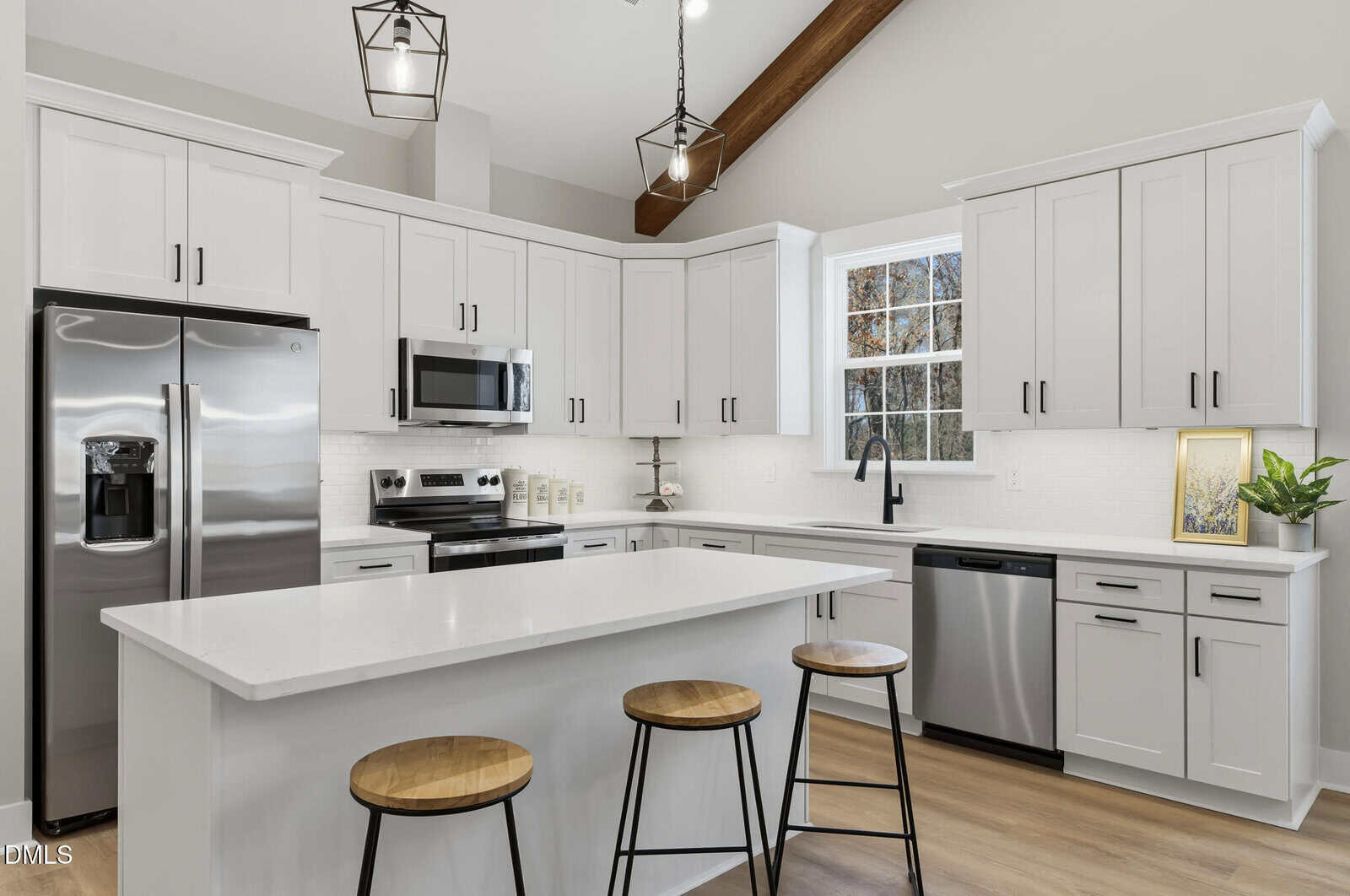 Modern kitchen with white cabinets, stainless steel appliances, a center island with three wooden stools, black fixtures, and pendant lights, featuring a window above the sink and neutral decor.
