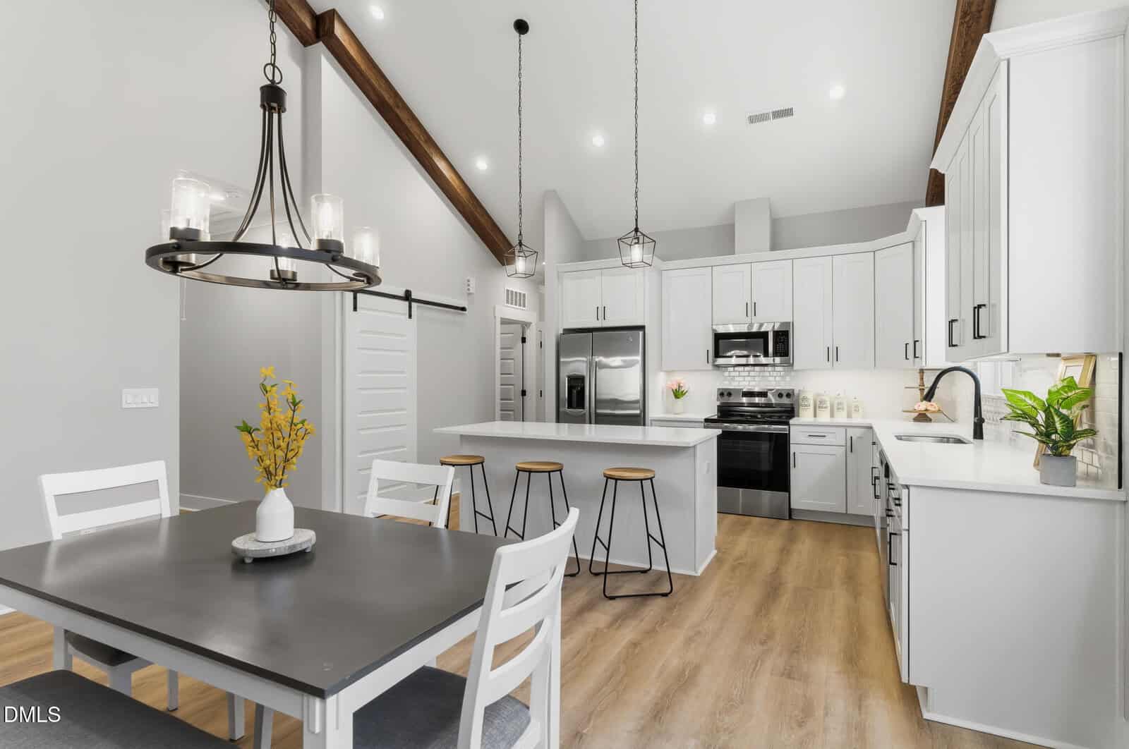 A modern kitchen and dining area with white cabinets, stainless steel appliances, a center island with three wooden stools, light wood flooring, pendant lighting, and a dining table with four chairs and a flower vase.