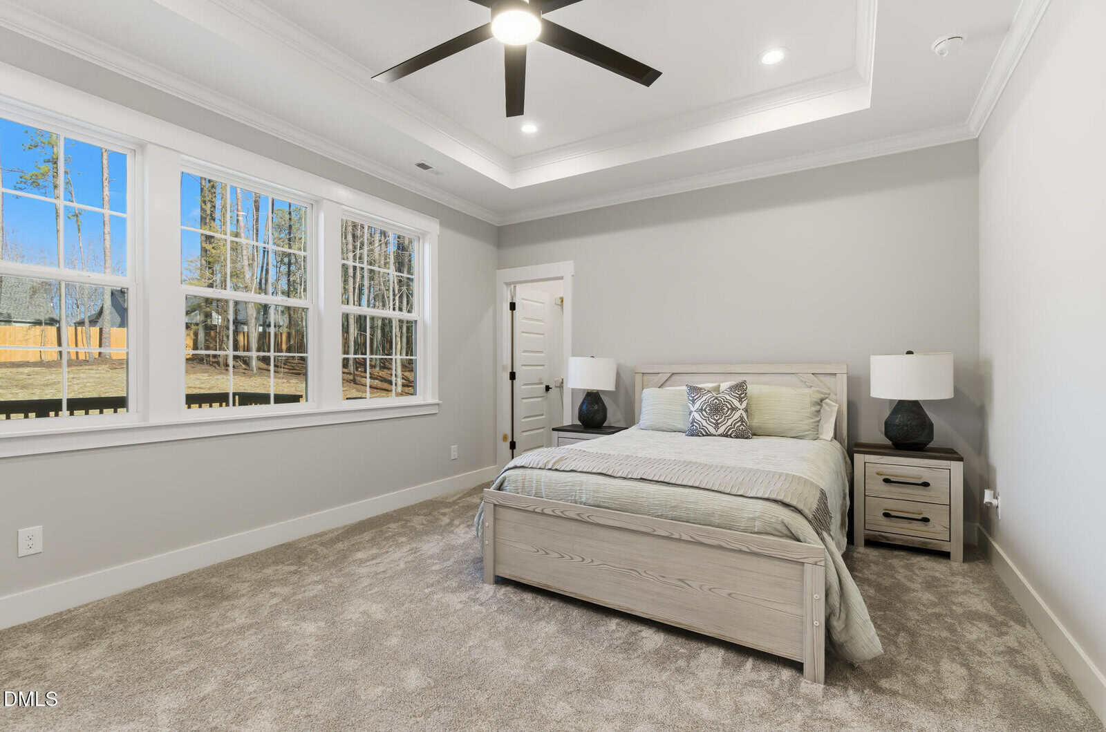 A neatly made bed with two nightstands and lamps sits in a carpeted bedroom with light gray walls. Large windows on the left let in natural light, and a ceiling fan is centered on the tray ceiling.
