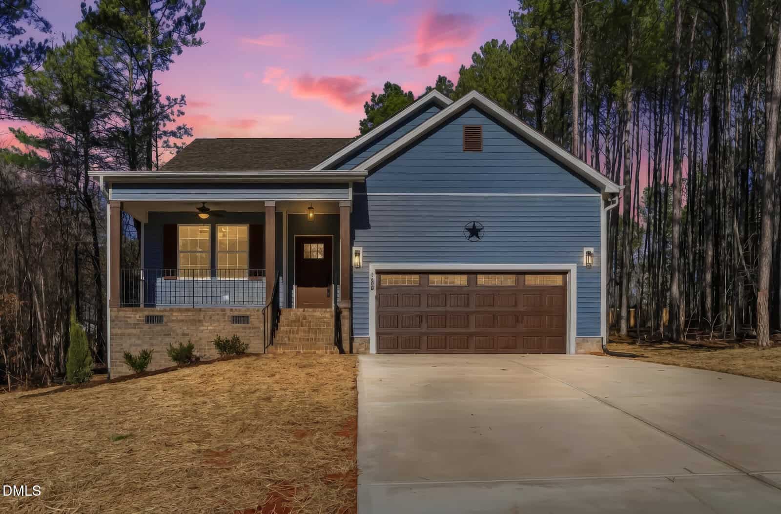 A blue single-story house at 128 Wichita Way in Louisburg features a front porch, brown double garage doors, and a concrete driveway, set against tall trees and a purple-pink sunset sky.