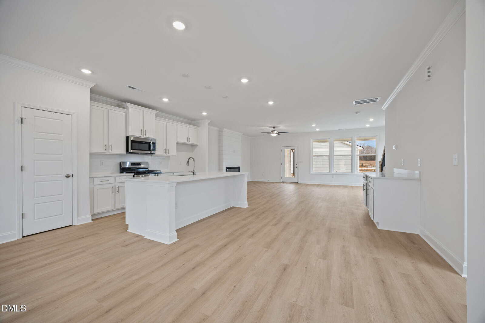 Spacious modern kitchen and living area with white cabinets, stainless steel appliances, a large island, light wood flooring, recessed lighting, and large windows letting in natural light.