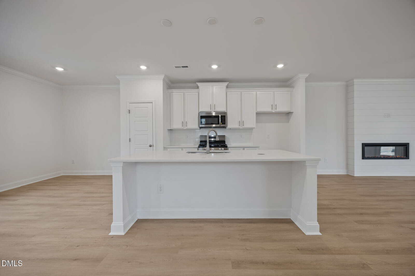 Modern kitchen with white cabinets, stainless steel appliances, a large island with a sink, light wood floors, and a built-in wall fireplace in a bright, open space.