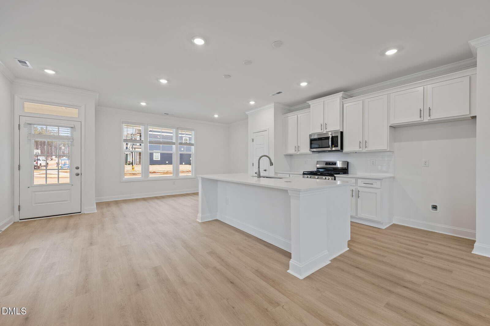 Open-concept kitchen and living area with light wood floors, white cabinets, a center island with sink, stainless steel appliances, and large front windows letting in natural light.