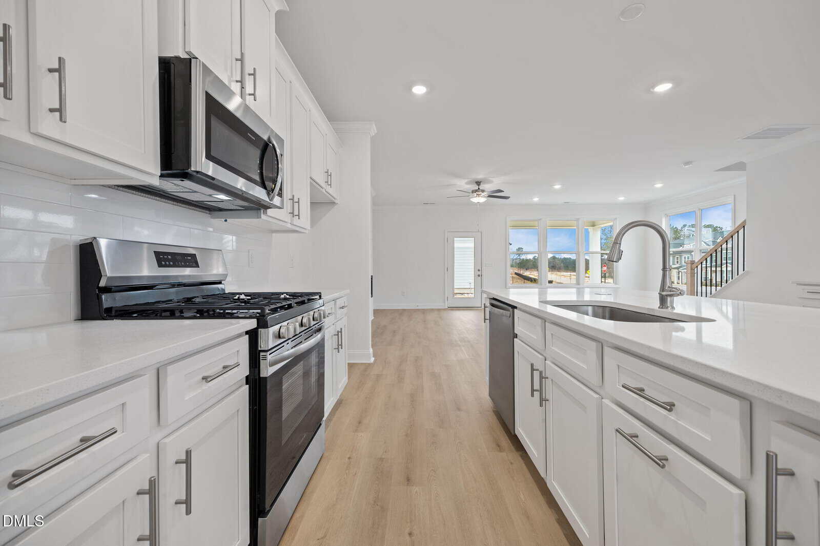 Modern, bright kitchen with white cabinets, stainless steel appliances, gas stove, built-in microwave, island with sink, and light wood flooring; open layout connects to a spacious living area with large windows and ceiling fan.
