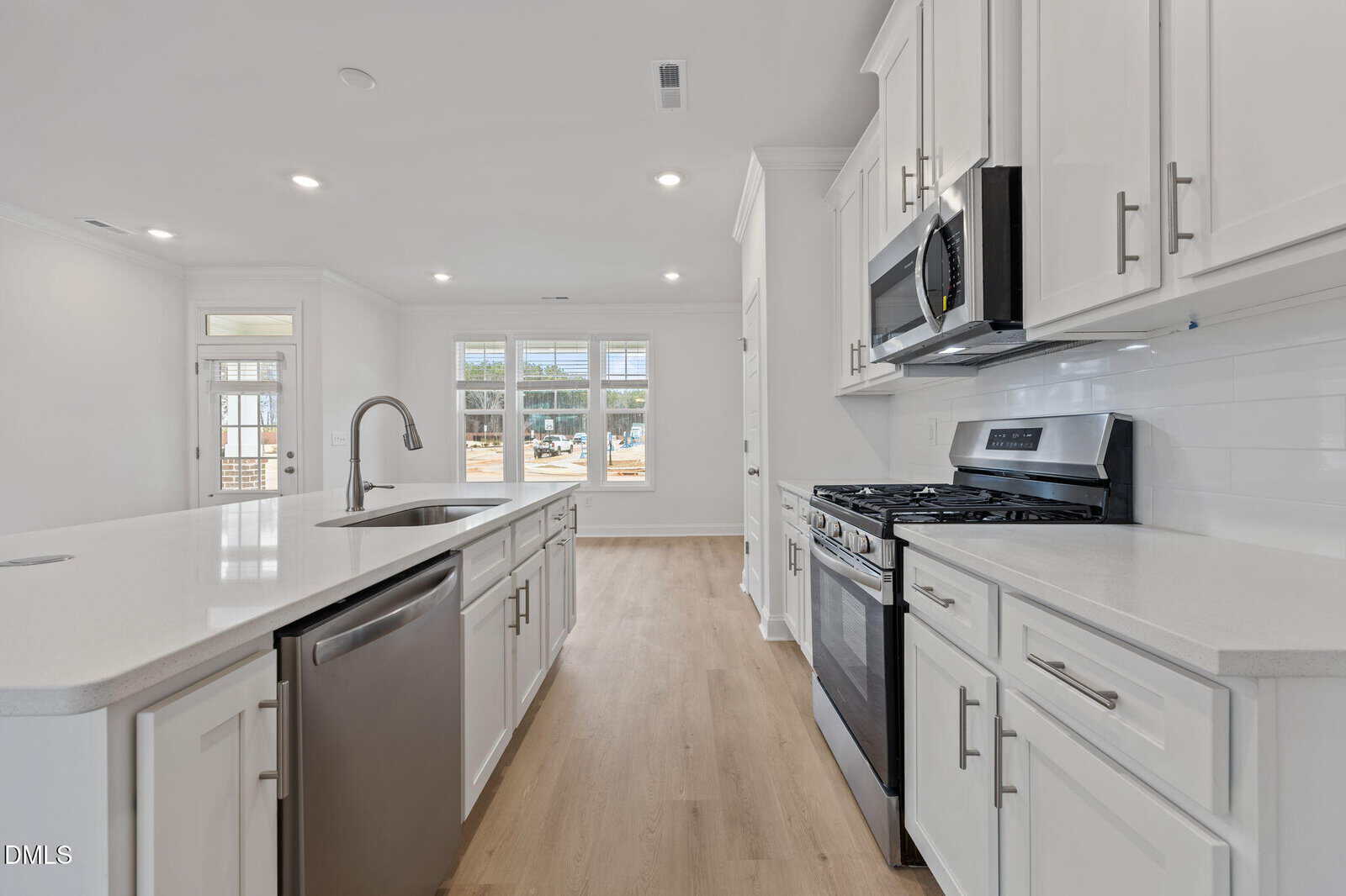 Modern kitchen with white cabinets, stainless steel appliances, a large island with sink and dishwasher, light wood flooring, and bright natural light from large windows and a glass door.