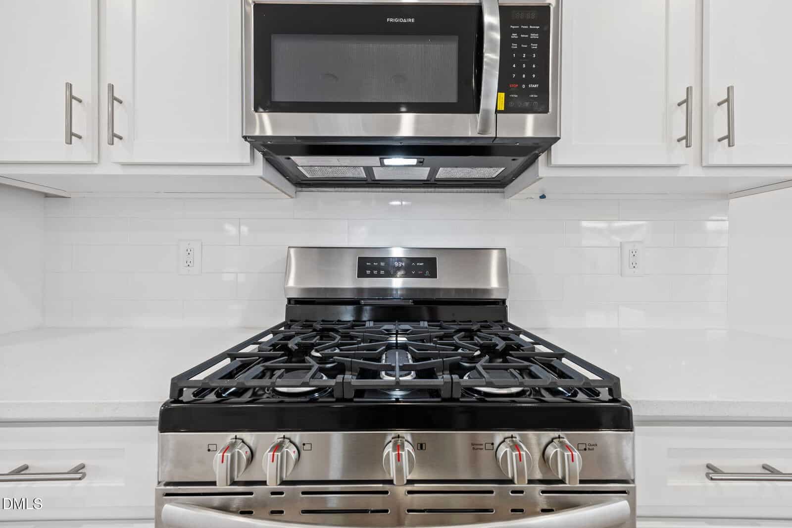 A modern kitchen features a stainless steel gas stove with four burners and control knobs, topped by a matching microwave oven, set against a white tile backsplash and white cabinetry.