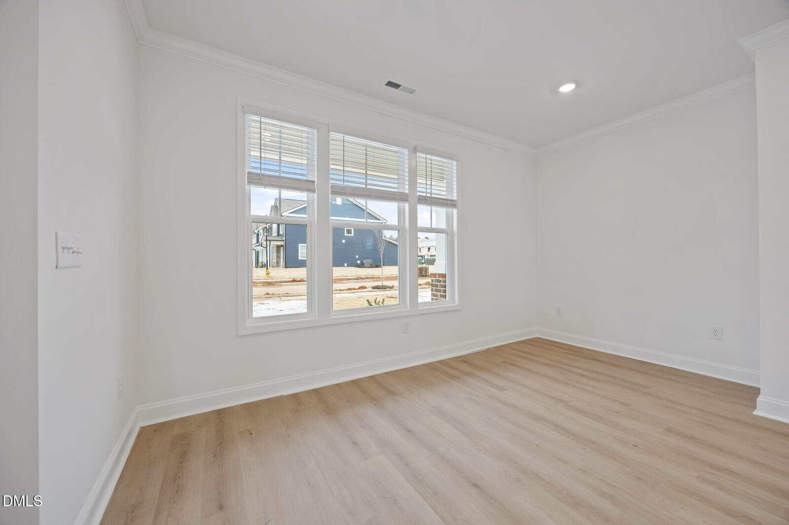 A bright, empty room with white walls, light wood flooring, and a large window letting in natural light. Suburban houses and a street are visible outside through the window.