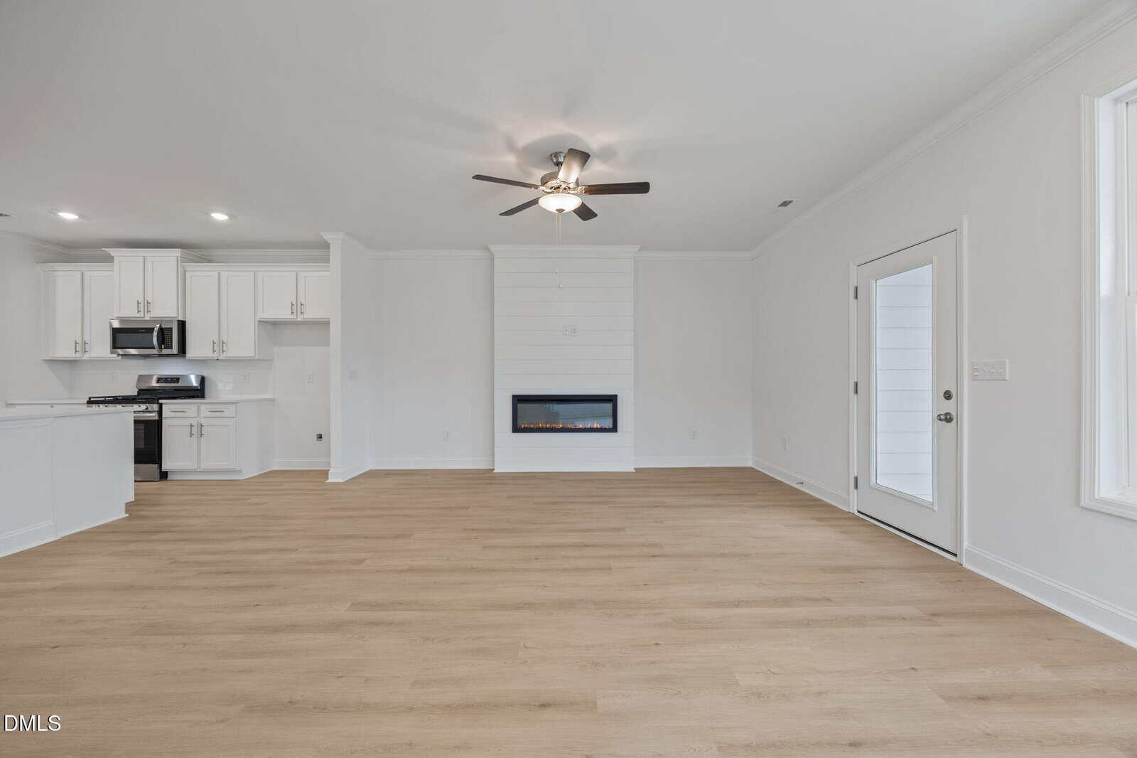 Modern, unfurnished living area with light wood floors, white walls, a ceiling fan, electric fireplace, and large windows. Kitchen with white cabinets and stainless steel appliances is visible to the left.