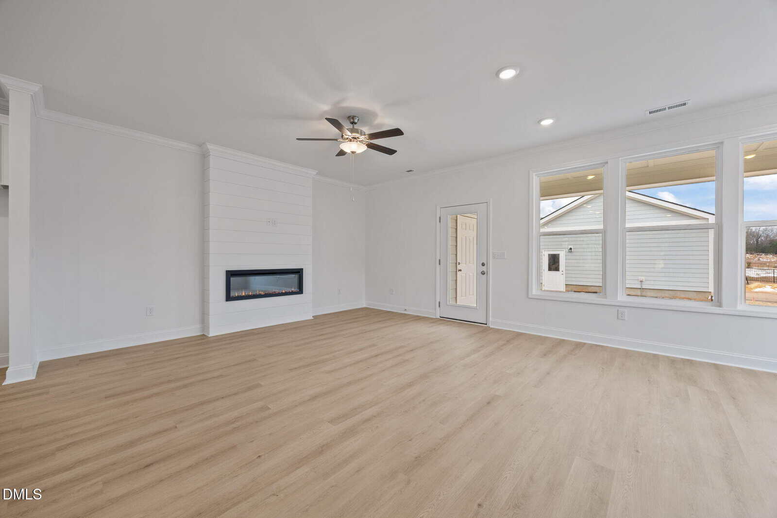 Spacious, empty living room with light wood flooring, white walls, a ceiling fan, built-in fireplace, and large windows letting in natural light. A door leads outside to a fenced yard.
