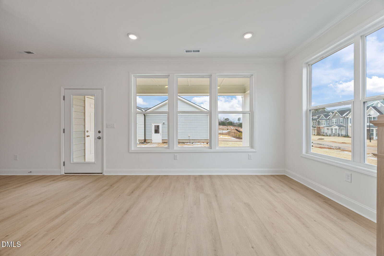 Bright, empty room with light wood floors, white walls, large windows, and a glass door. Outside, other houses are visible under a partly cloudy sky.