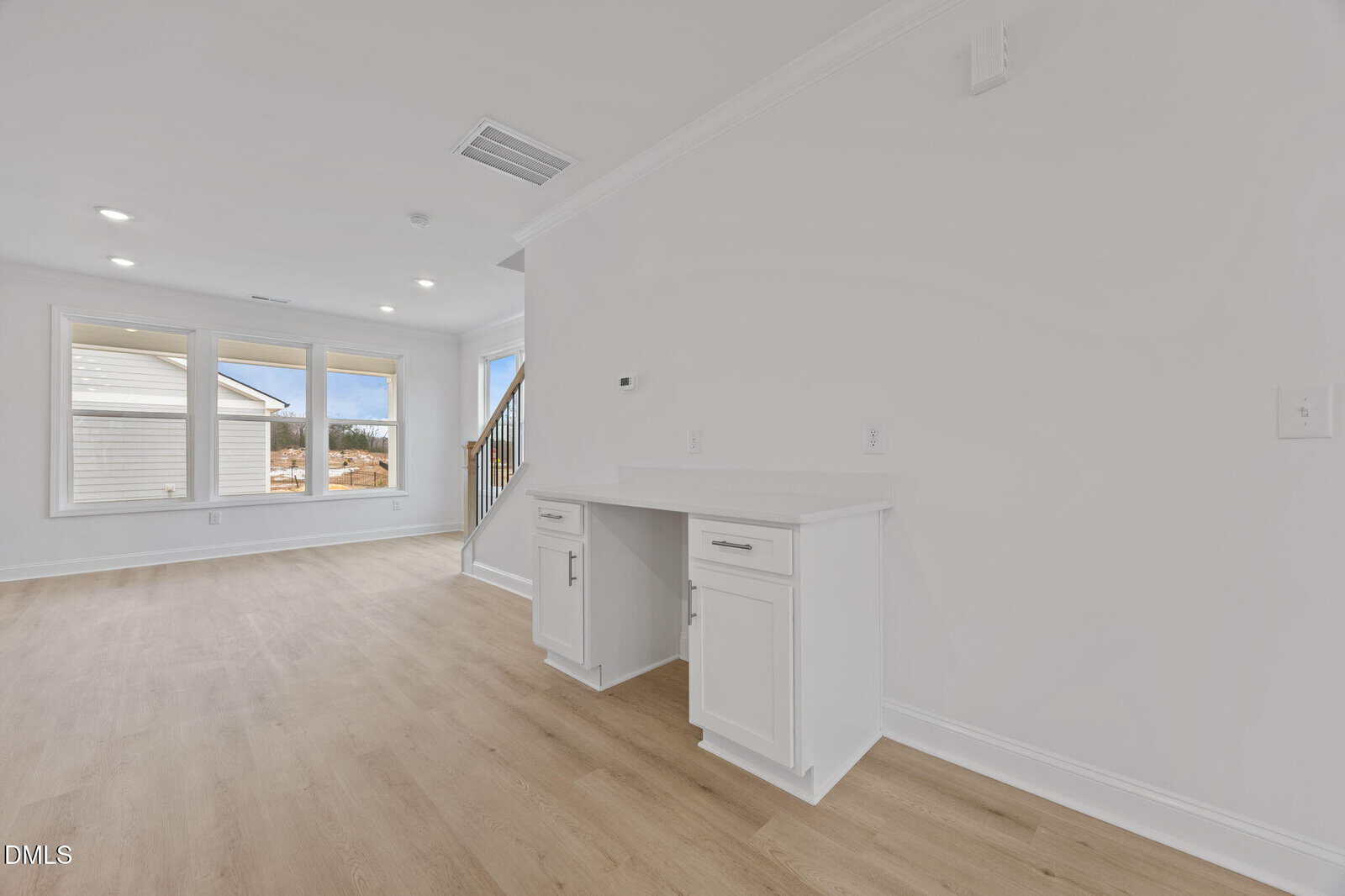 Bright, modern room with light wood floors, white walls, large windows, and a built-in white desk with cabinets beneath. A staircase and outdoor view are visible in the background.