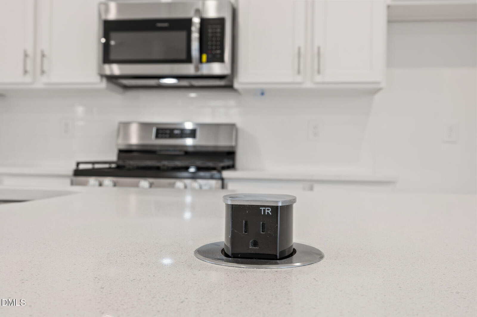 A pop-up electrical outlet and USB port are extended from a white kitchen island countertop, with a stove, oven, microwave, and white cabinets in the background.