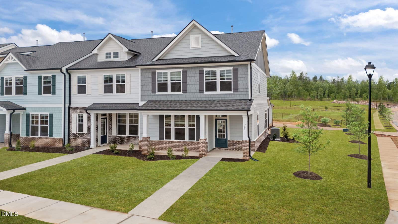 A modern two-story townhouse with gray siding, white trim, and brick accents, featuring a small front porch, manicured lawn, and a sidewalk leading to the entrance. A streetlamp and trees line the sidewalk.