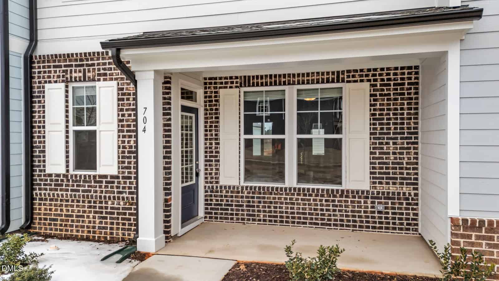 Front porch of a house with a covered entrance, black door with glass panel, large window with white shutters, brick and white siding exterior, and small bushes by the walkway. House number 704 is visible on the porch column.