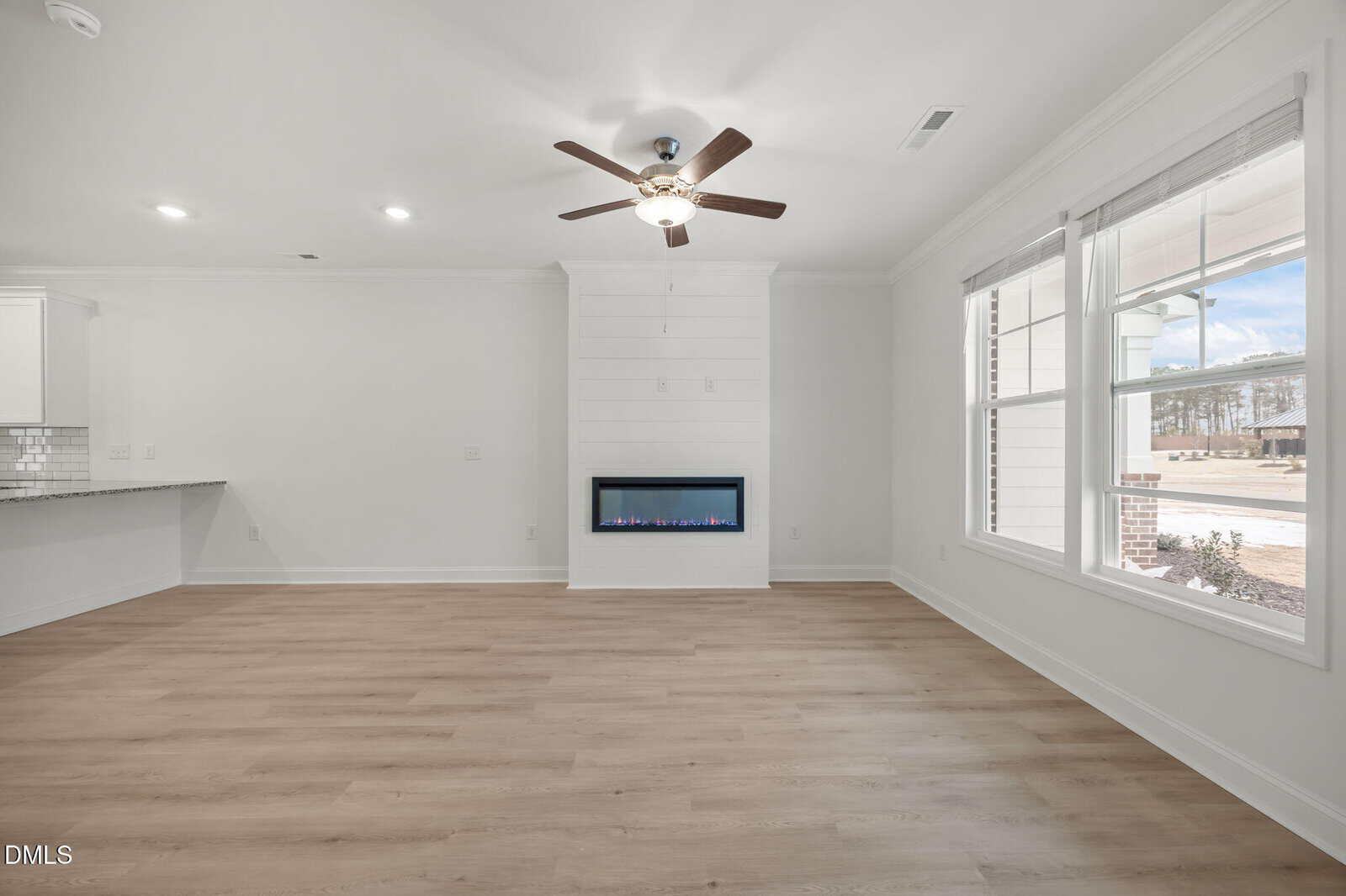 A bright, modern living room with light wood floors, a ceiling fan, built-in electric fireplace, and large windows letting in natural light. White walls and an open layout create a spacious feel.