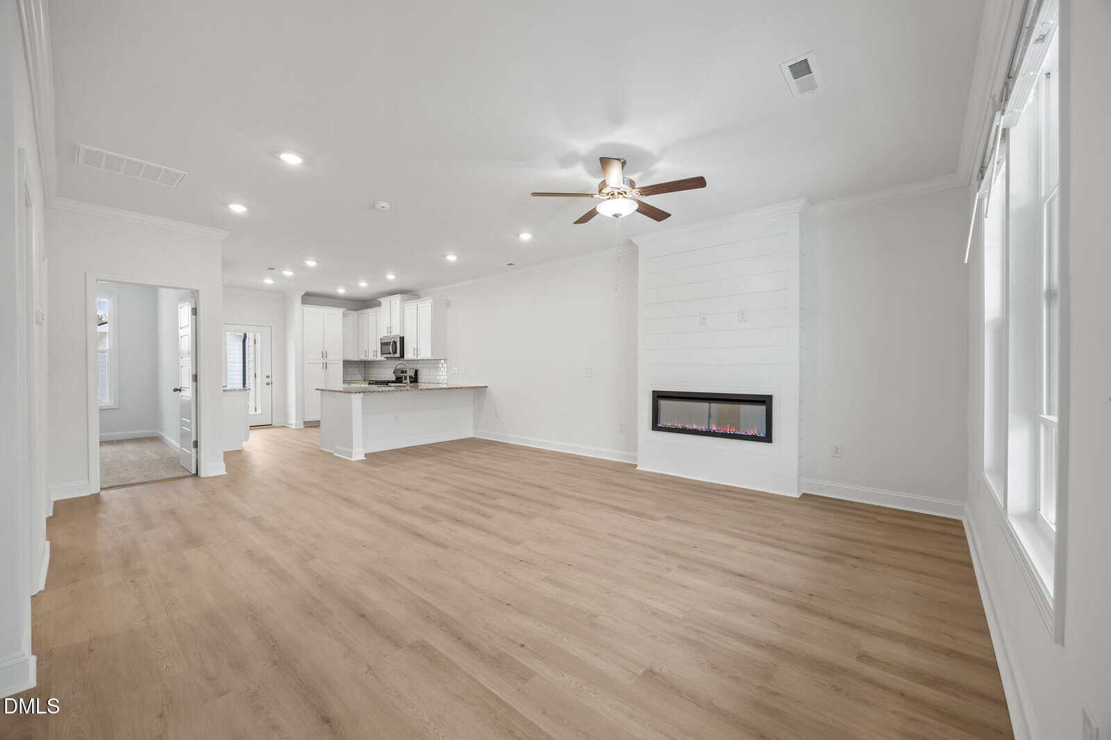 Bright open living area with wood flooring, white walls, a ceiling fan, recessed lighting, and an electric fireplace. The space flows into a modern kitchen with white cabinets and stainless steel appliances.