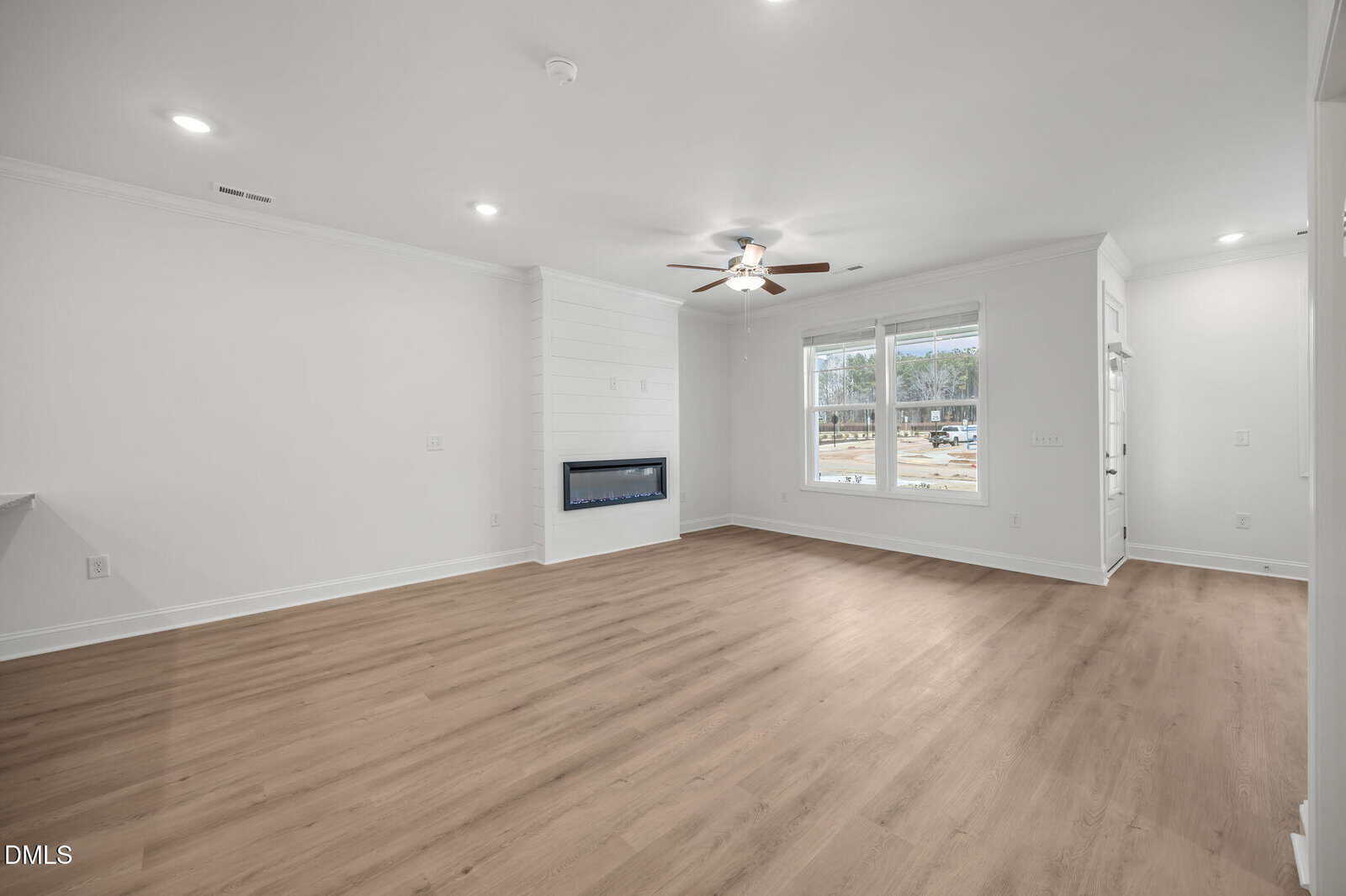 A bright, empty living room with light wood flooring, white walls, a modern electric fireplace, ceiling fan, and large windows allowing natural light to fill the space.