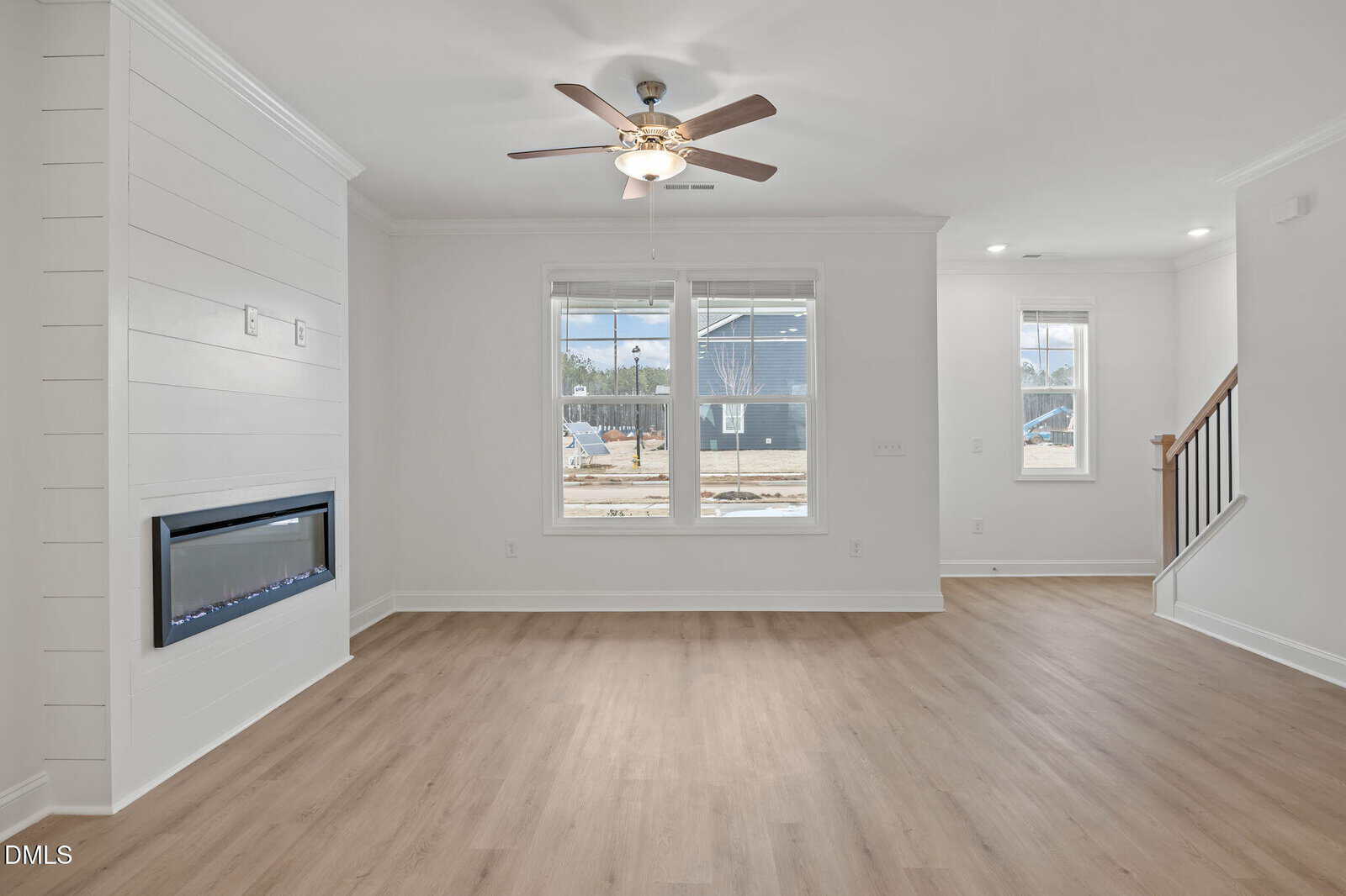 Bright living room with light wood flooring, white walls, ceiling fan with light, built-in electric fireplace, large windows, and a partial view of a staircase and entryway.