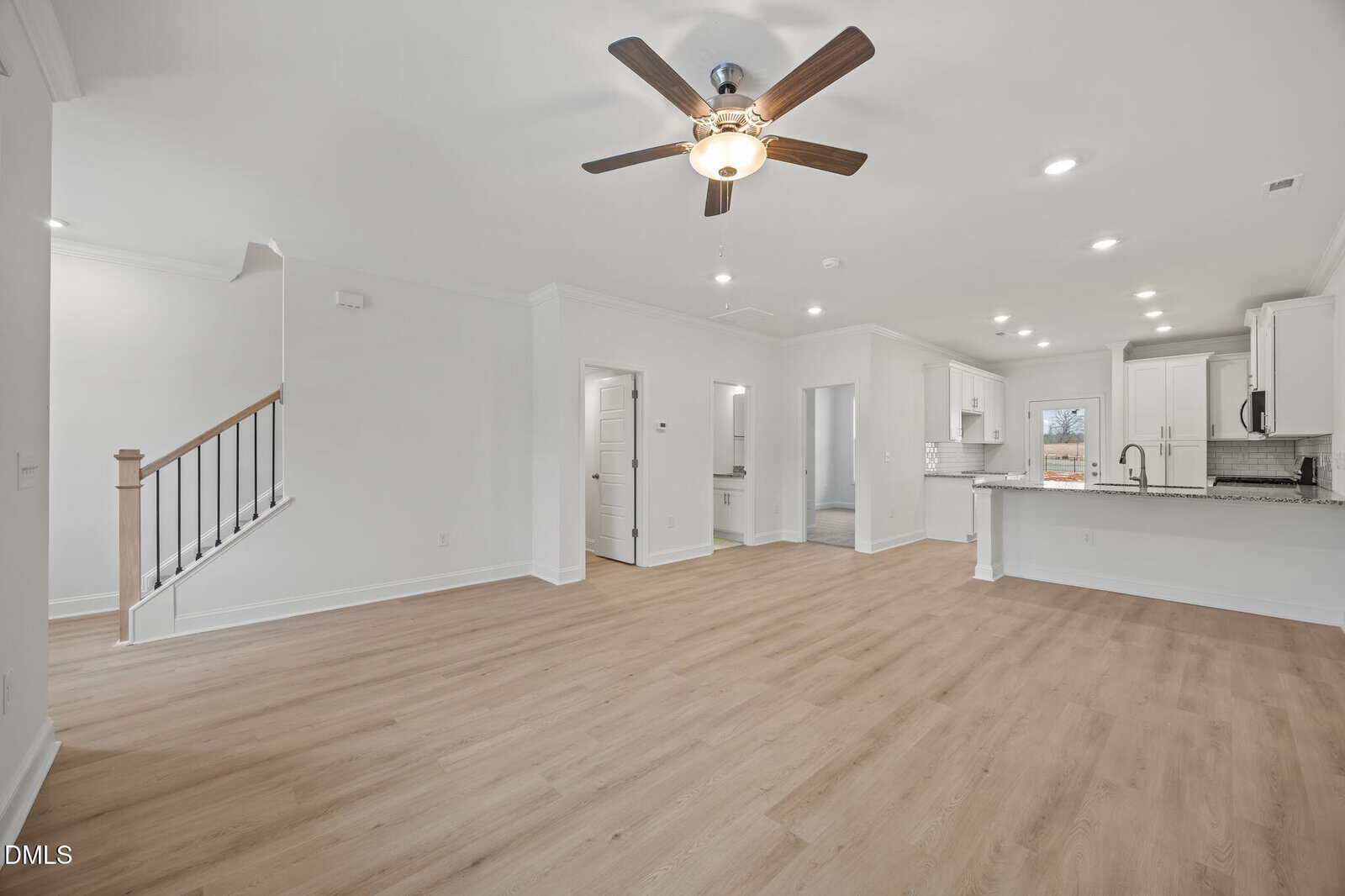 Bright, empty open-concept living area with light wood floors, ceiling fan, white walls, staircase, recessed lighting, and a modern kitchen with white cabinets and granite countertops in the background.