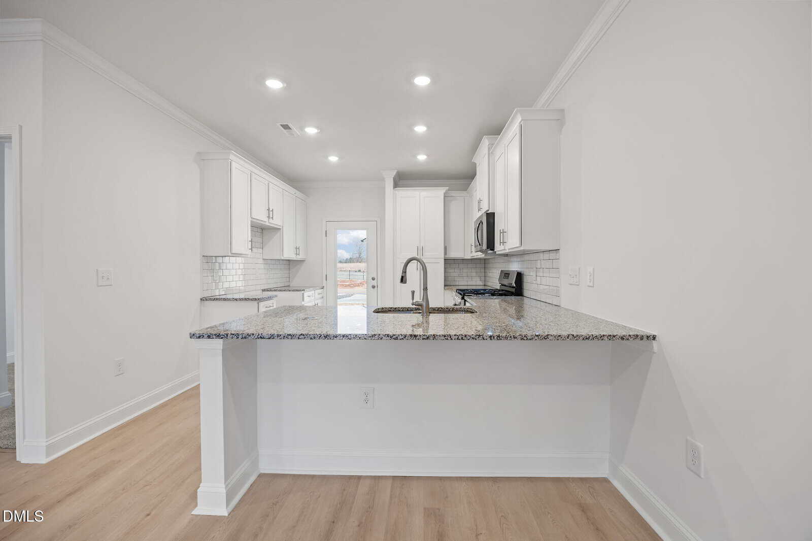 Modern kitchen with white cabinets, granite countertops, subway tile backsplash, stainless steel appliances, under-cabinet lighting, and light wood flooring, with a breakfast bar and a door leading outside in the background.