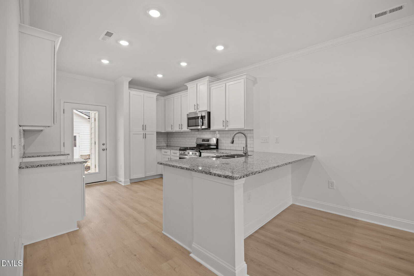 Modern kitchen with white cabinets, stainless steel appliances, granite countertops, and light wood flooring. Recessed lighting brightens the space, and a glass door leads outside. The design is clean and open.