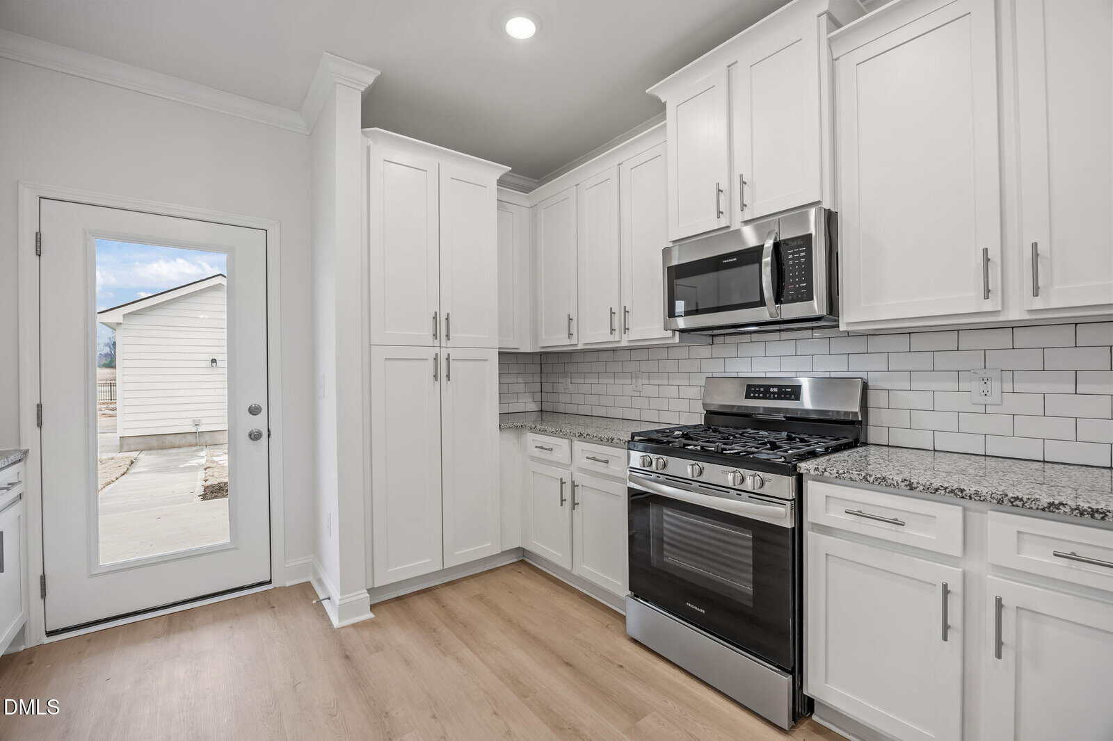 Modern kitchen with white cabinets, stainless steel appliances, granite countertops, subway tile backsplash, and light wood flooring. A glass door leads outside, bringing in natural light.