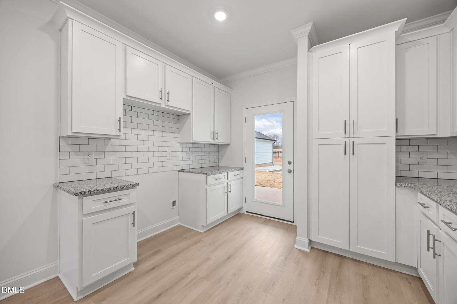 Bright, modern kitchen with white cabinets, white subway tile backsplash, granite countertops, light wood flooring, and a glass door leading outside to a yard.