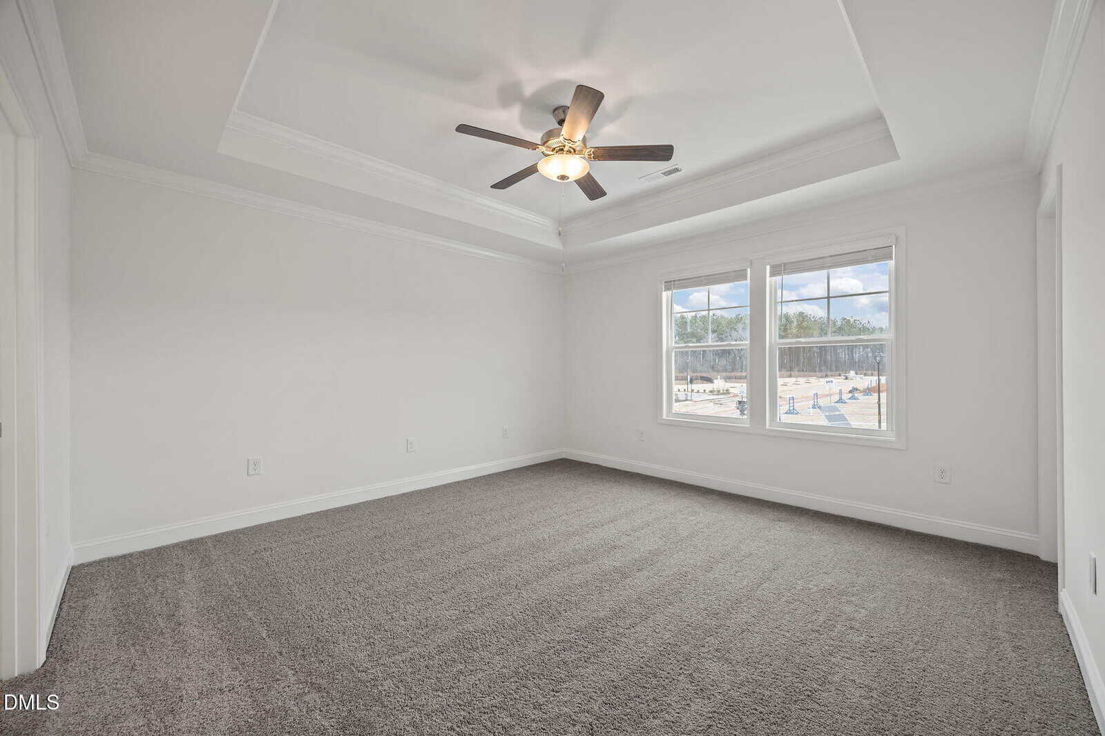 Empty, bright room with white walls, gray carpet, tray ceiling, and ceiling fan. Two large windows let in natural light and offer a view of a construction site outside.