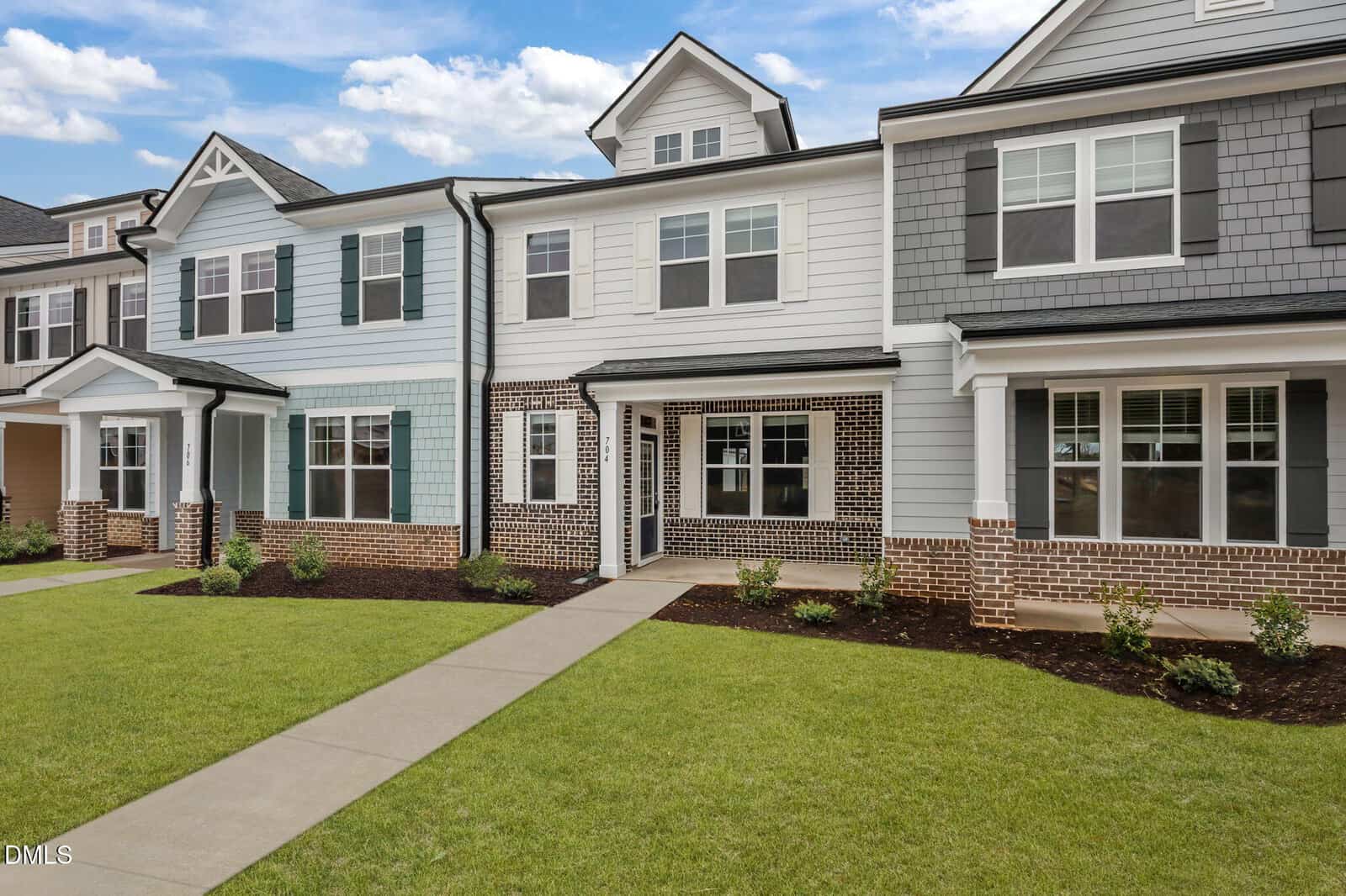 Modern two-story townhouse with light blue and gray siding, white trim, brick accents, large front windows, a covered porch, manicured lawn, and a sidewalk leading to the front entrance.