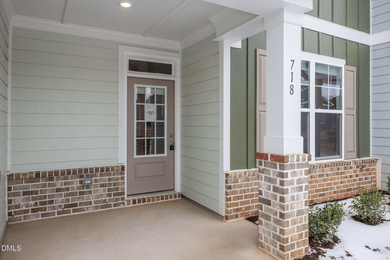 Covered front porch of a house with light green siding, white trim, and brick accents. The house number 718 is displayed on a white column next to a glass-paneled front door. Small shrubs line the edge of the porch.