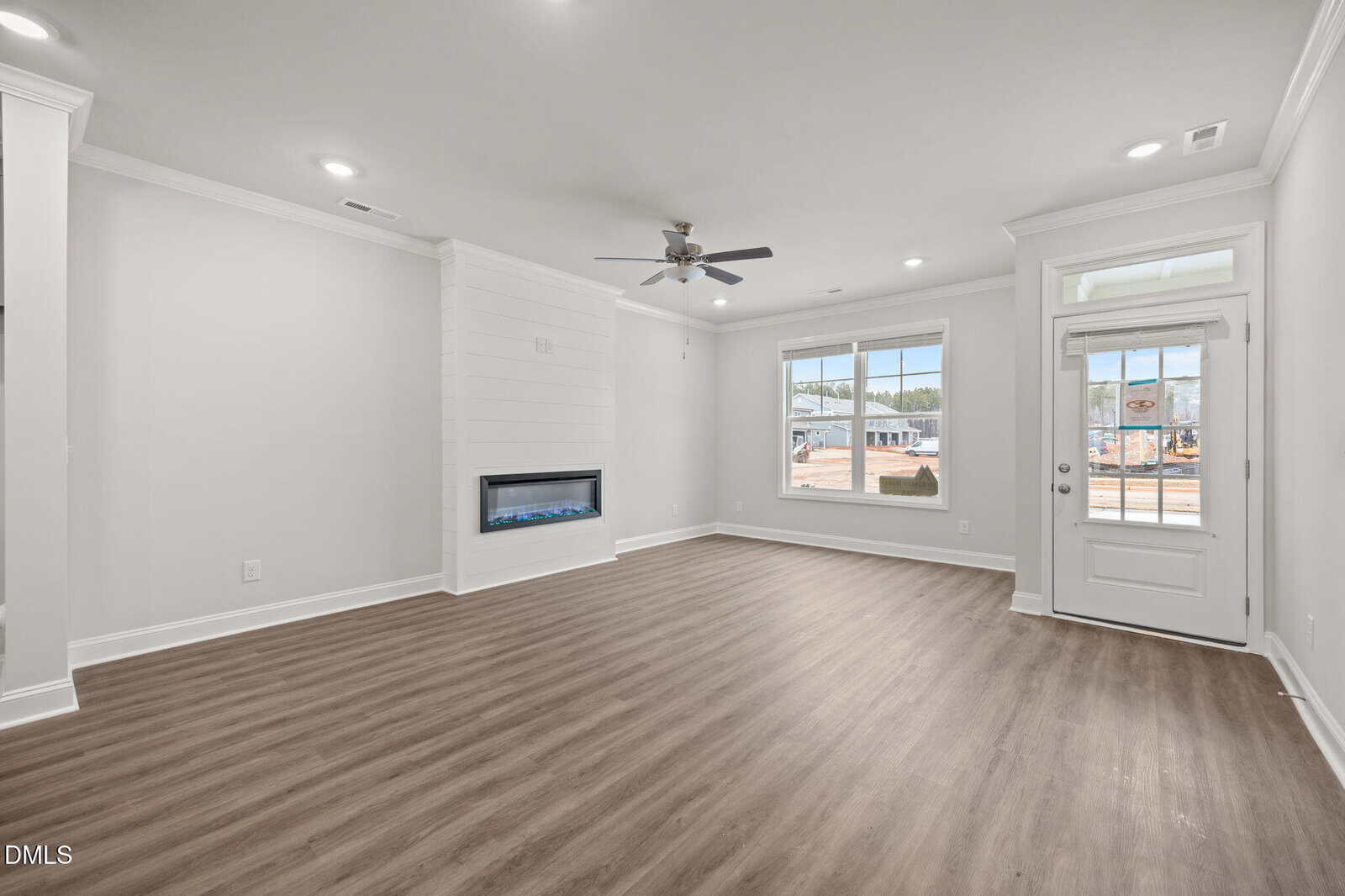 A spacious, empty living room with light gray walls, wood-style flooring, a ceiling fan, large windows, and a glass door. A modern electric fireplace is built into a white shiplap accent wall.