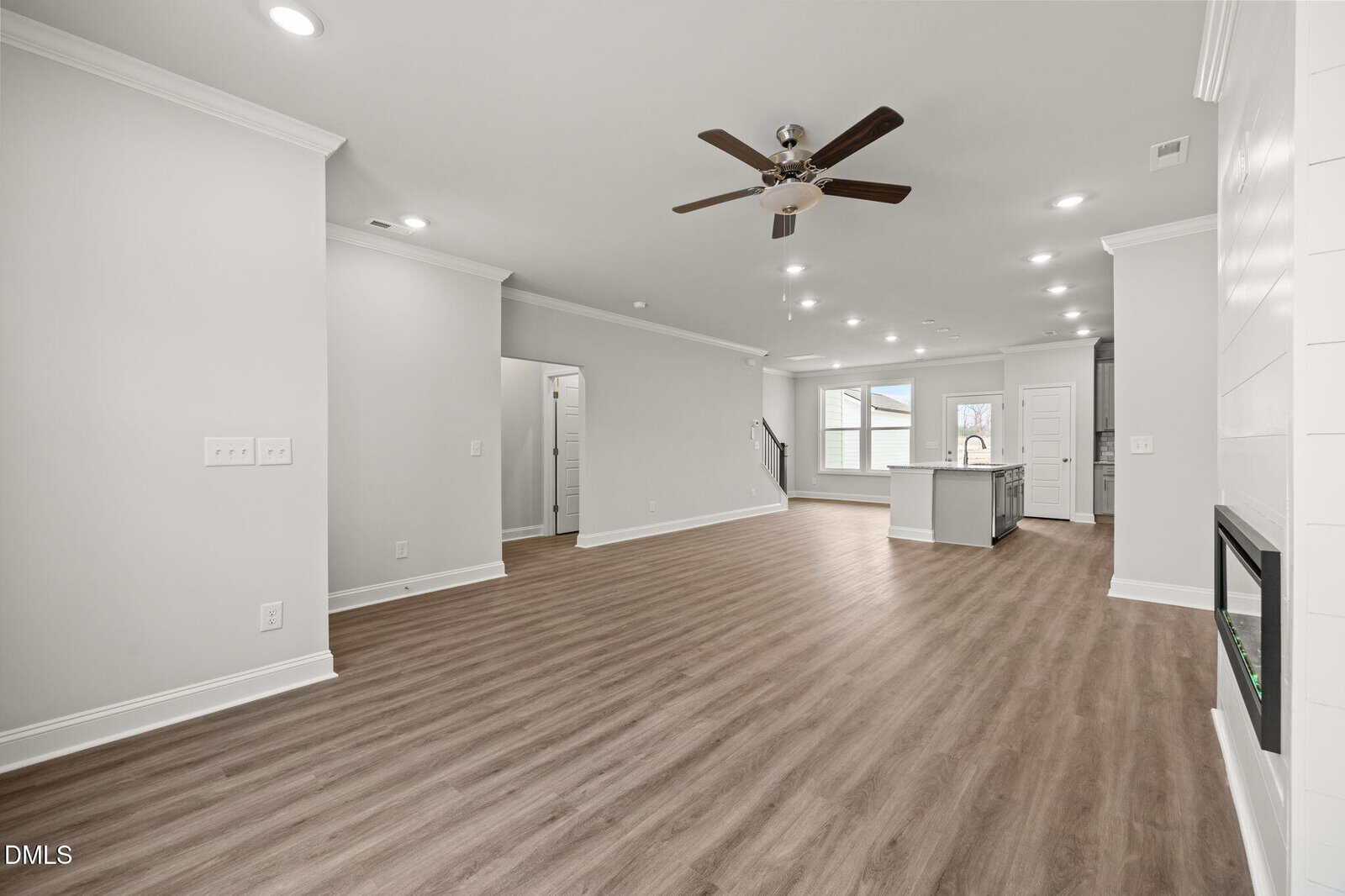 Spacious, modern open-concept living area with light gray walls, wood flooring, ceiling fan, recessed lighting, a fireplace, and a kitchen with an island and large windows in the background.