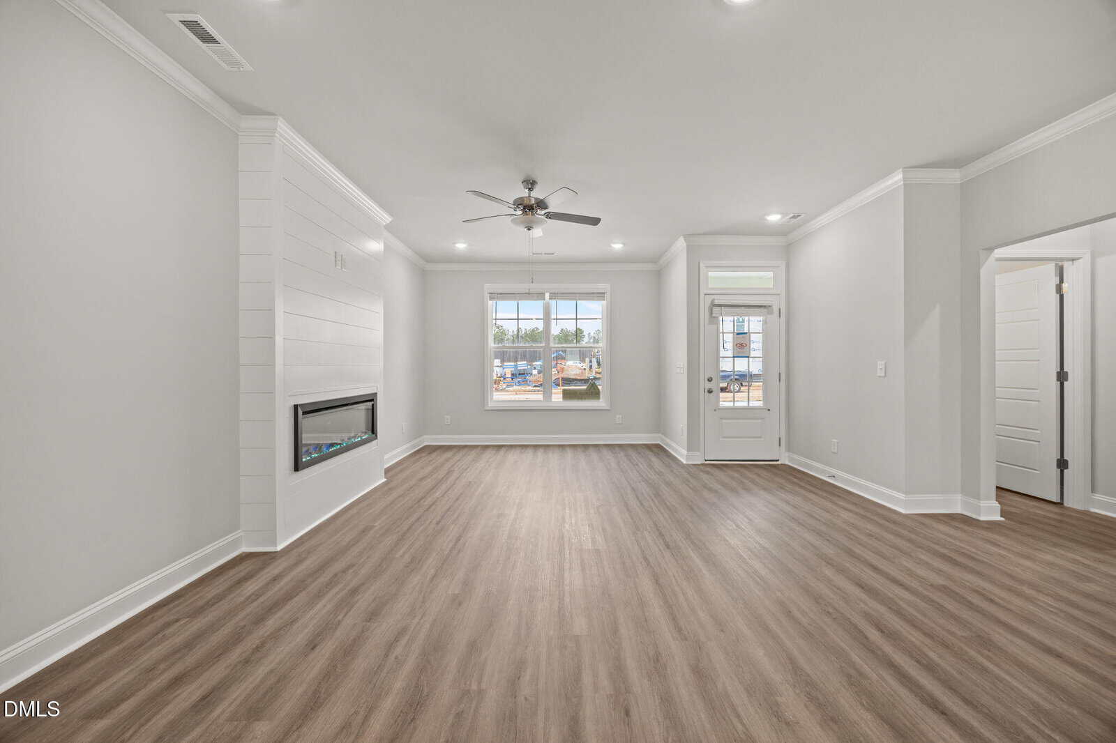 Bright, empty living room with light wood flooring, white walls, a ceiling fan, built-in fireplace, large front window, and a glass door leading outside. Modern, minimal design with lots of natural light.