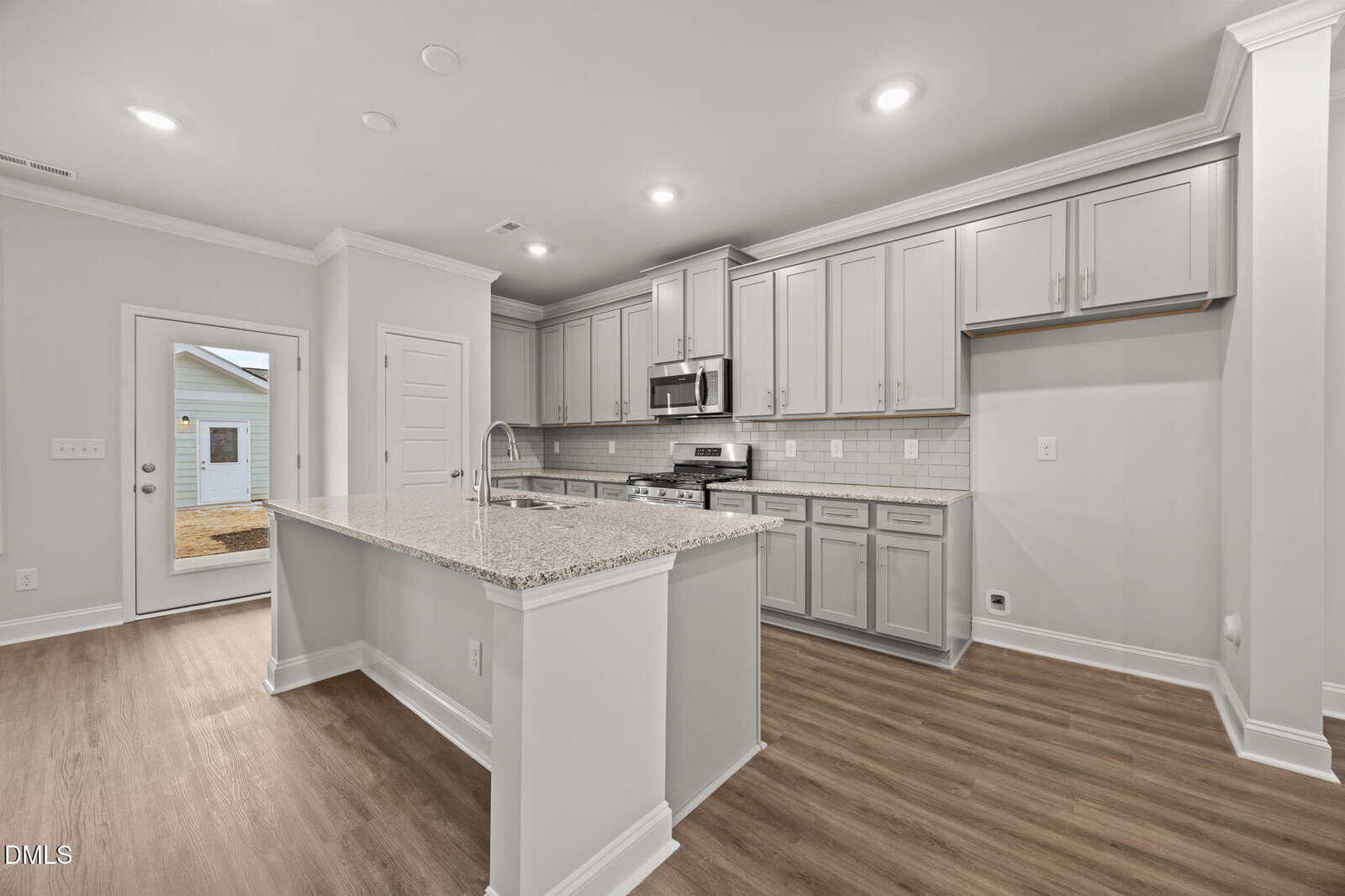 Modern kitchen with light gray cabinets, stainless steel appliances, a large granite island, subway tile backsplash, and wood-style flooring. A glass door leads outside, and recessed ceiling lights brighten the space.