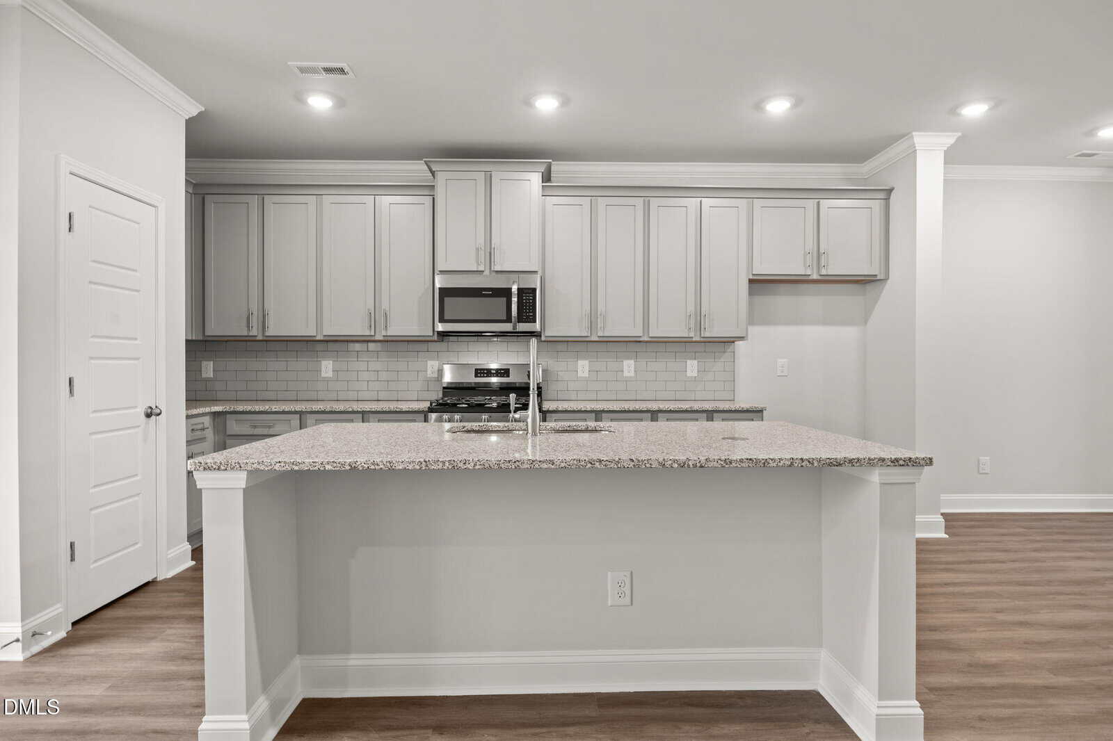 Modern kitchen with light gray cabinets, stainless steel appliances, a built-in microwave, white subway tile backsplash, and a large granite island countertop; wood flooring and recessed ceiling lights are visible.