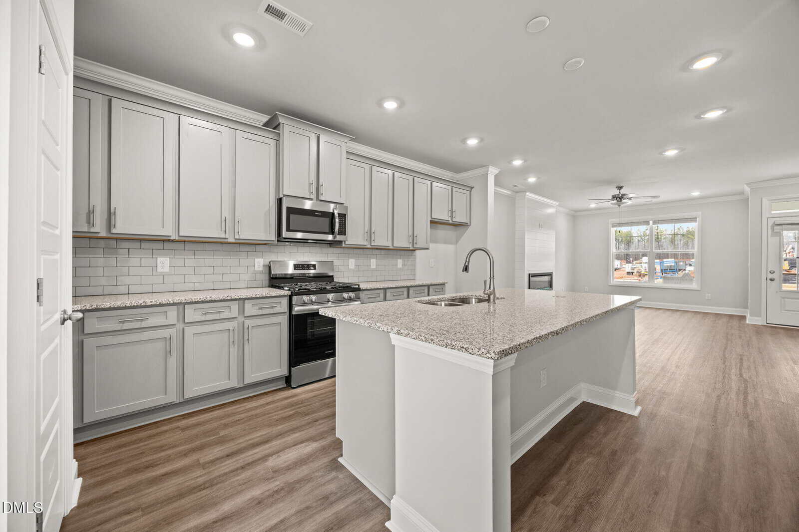 Modern kitchen with light gray cabinets, stainless steel appliances, white subway tile backsplash, large island with sink, and wood-look flooring, open to a bright living area with large windows and ceiling fan.