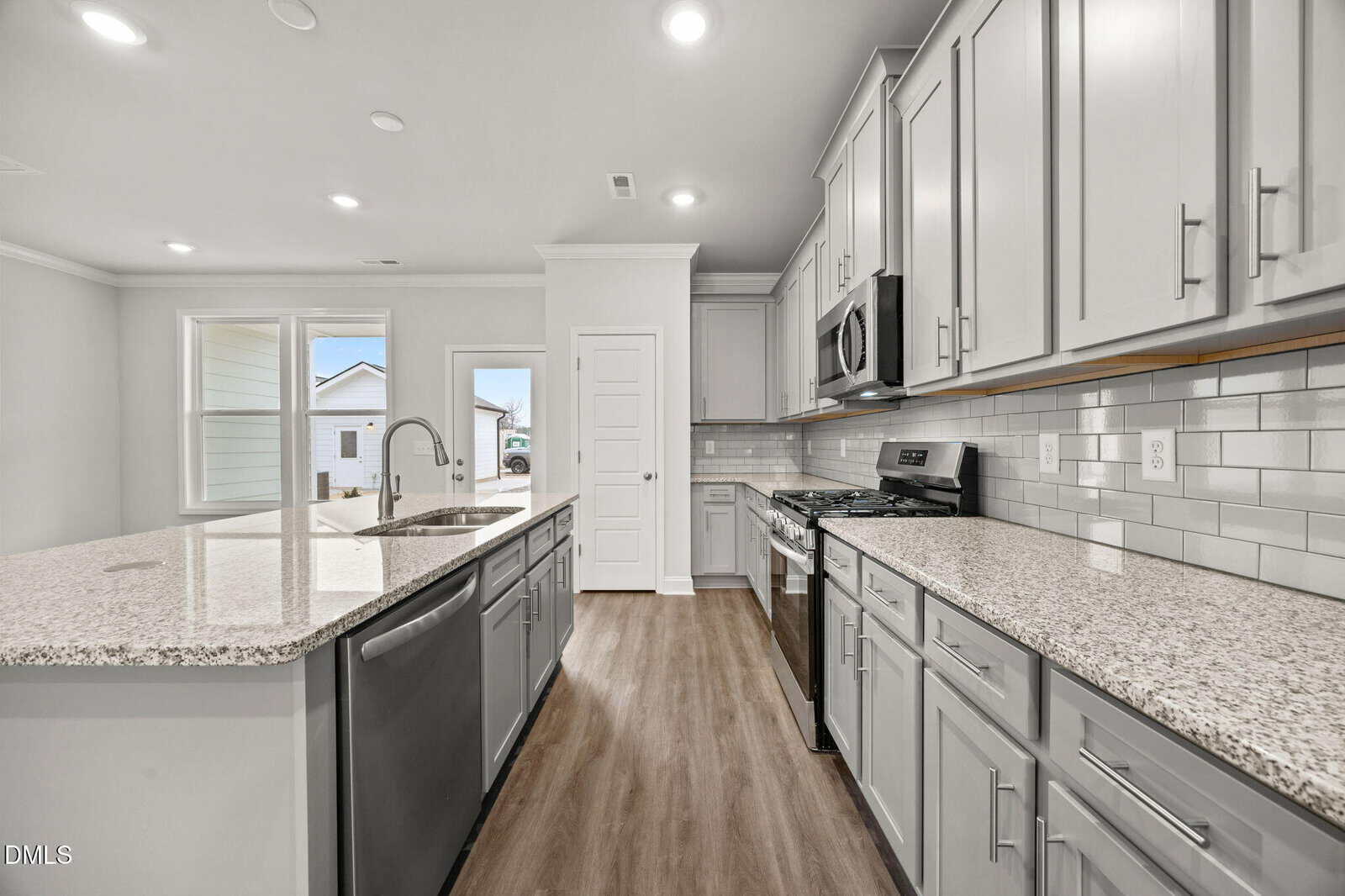 A modern kitchen with light gray cabinets, stainless steel appliances, granite countertops, a center island with a sink, subway tile backsplash, and wood-look flooring. Large windows and a door provide natural light.