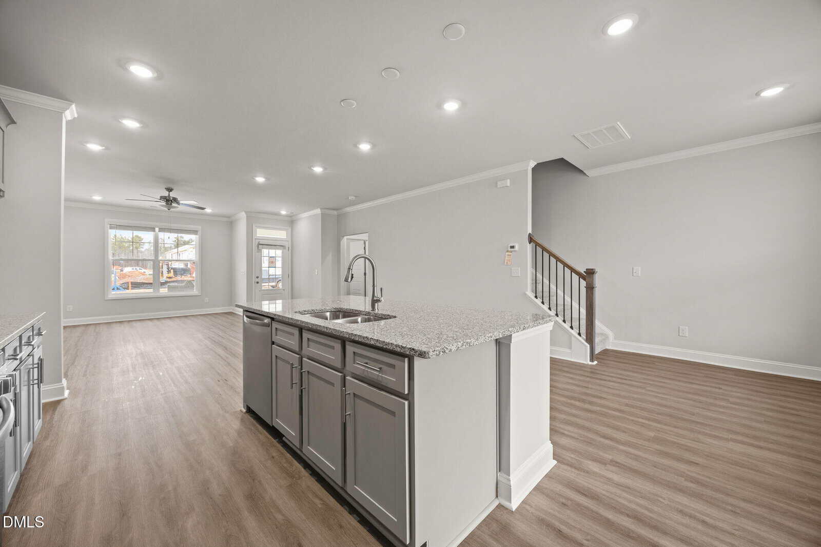 Modern, open-concept kitchen and living area with light gray walls, wood flooring, recessed lighting, a central island with a sink, and a staircase leading to the upper floor. Large windows provide natural light.