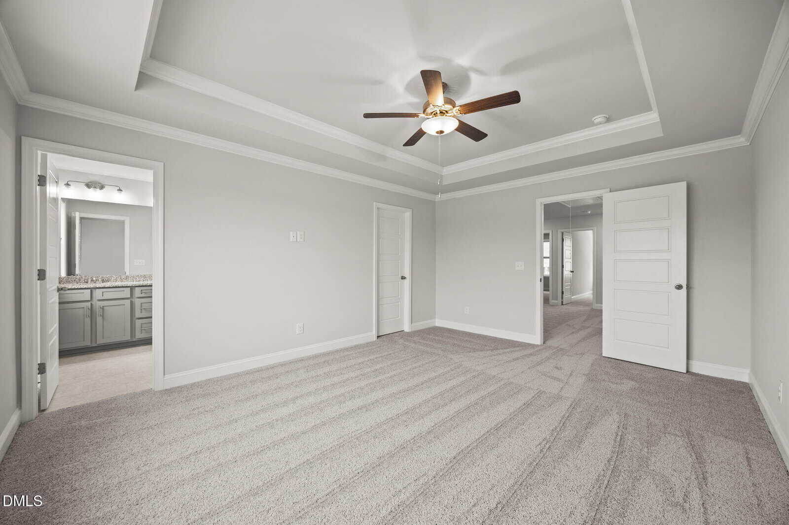Spacious, empty bedroom with light gray walls, white trim, tray ceiling, ceiling fan, and carpeted floor. Two open doors reveal a bathroom with double sinks and a hallway.