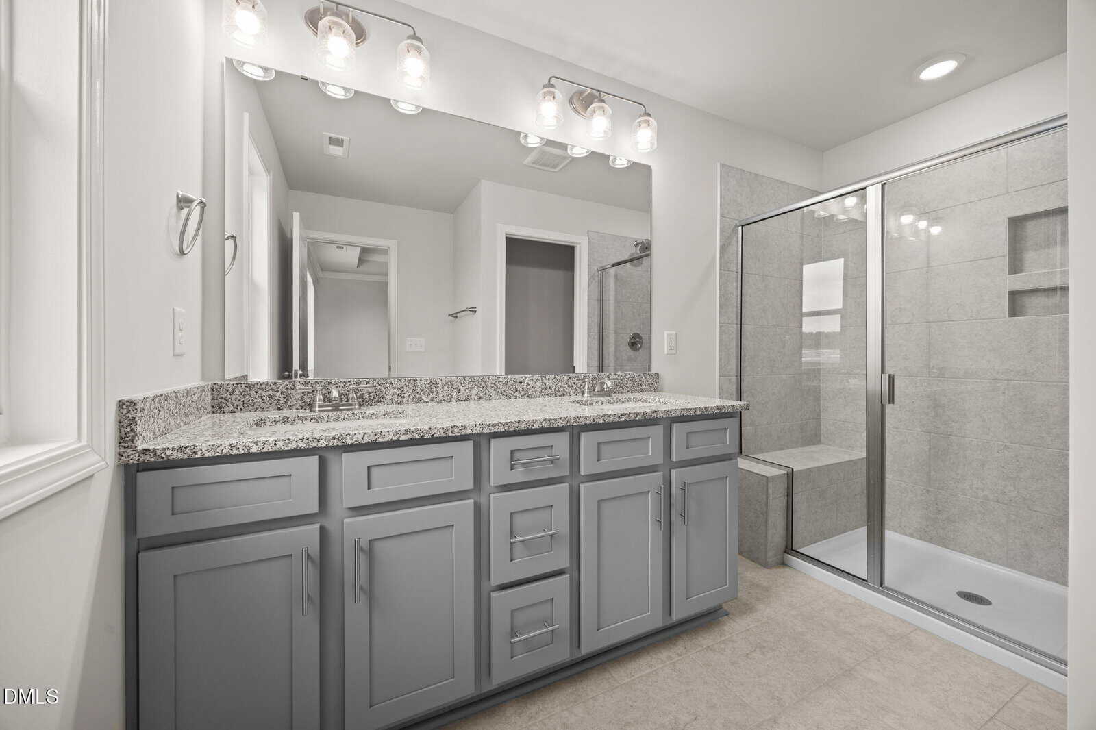 Modern bathroom with a large mirror, double sink vanity with gray cabinets, granite countertop, glass-enclosed shower with built-in bench, and neutral tile flooring. Bright lighting and clean design.