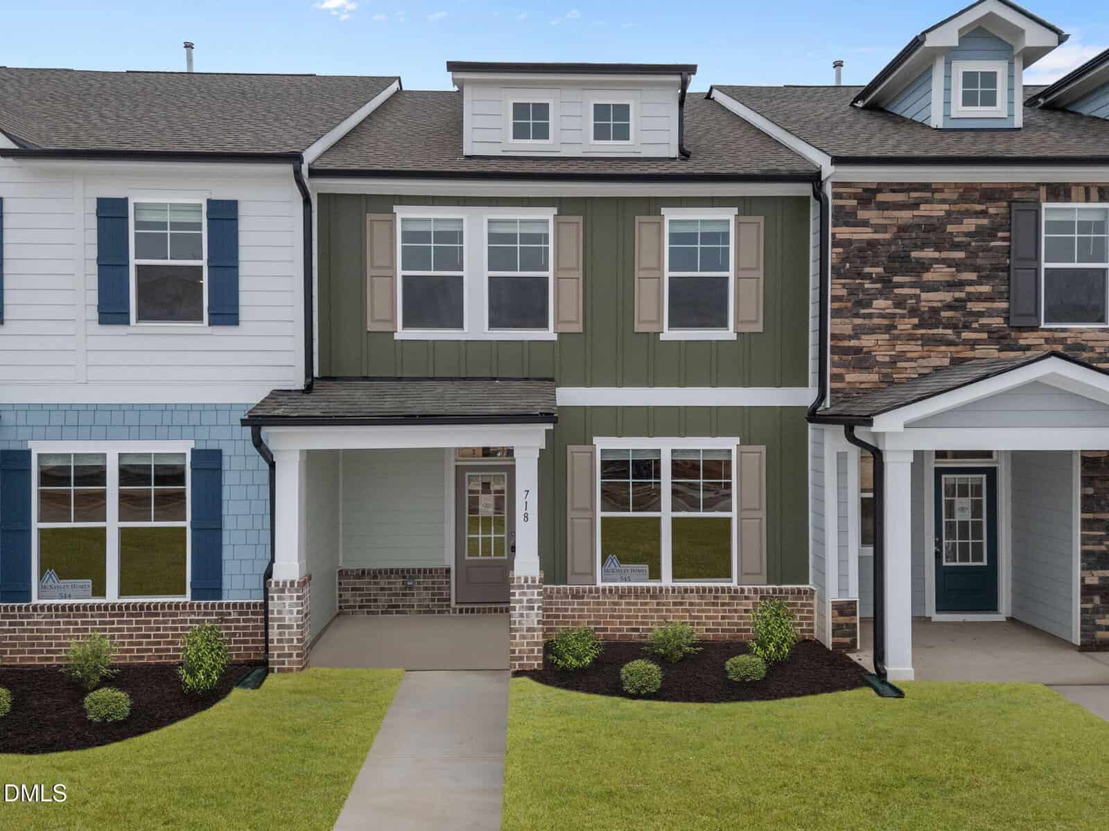 A modern two-story townhouse with green and white siding, brick accents, and a covered front porch, surrounded by small shrubs and mulch, between two similar homes.