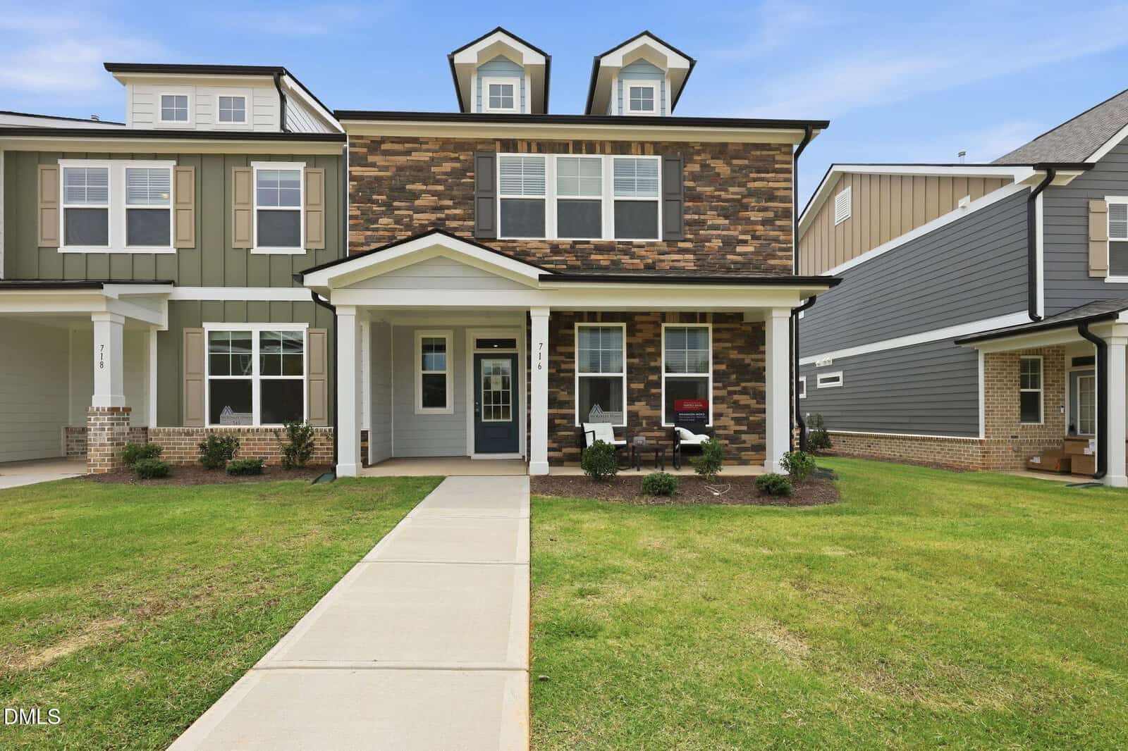 Two-story suburban house with mixed stone and siding exterior, front porch with chairs, manicured lawn, and a sidewalk leading to the front door. Neighboring houses are visible on either side.