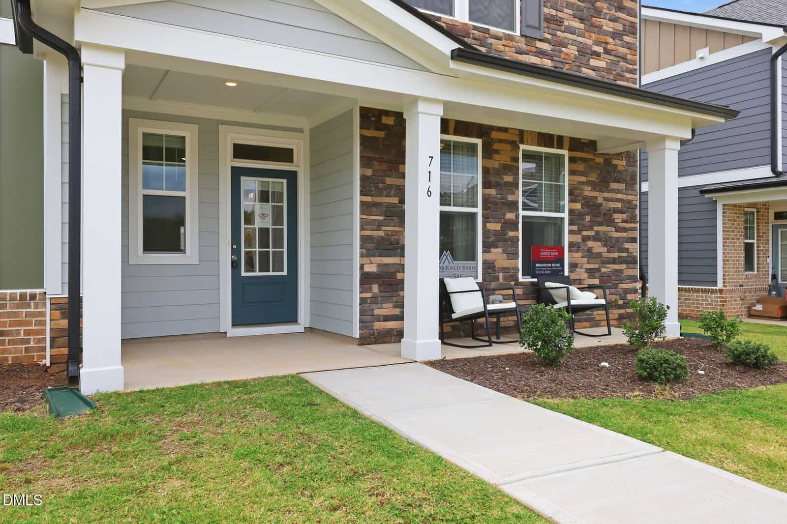 Modern house with stone and gray siding, covered front porch, white pillars, blue door, and two black chairs with white cushions. Green lawn and small shrubs line the walkway.