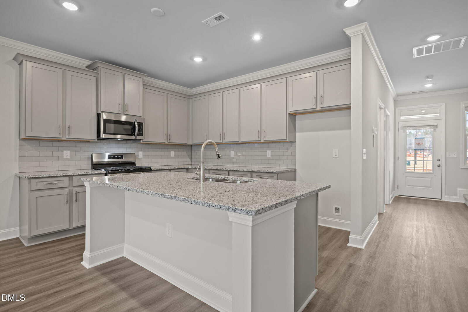 Modern kitchen with light gray cabinets, stainless steel appliances, a large center island with a sink, granite countertops, subway tile backsplash, and wood-look flooring. Bright lighting and a hallway leading to a door are visible.
