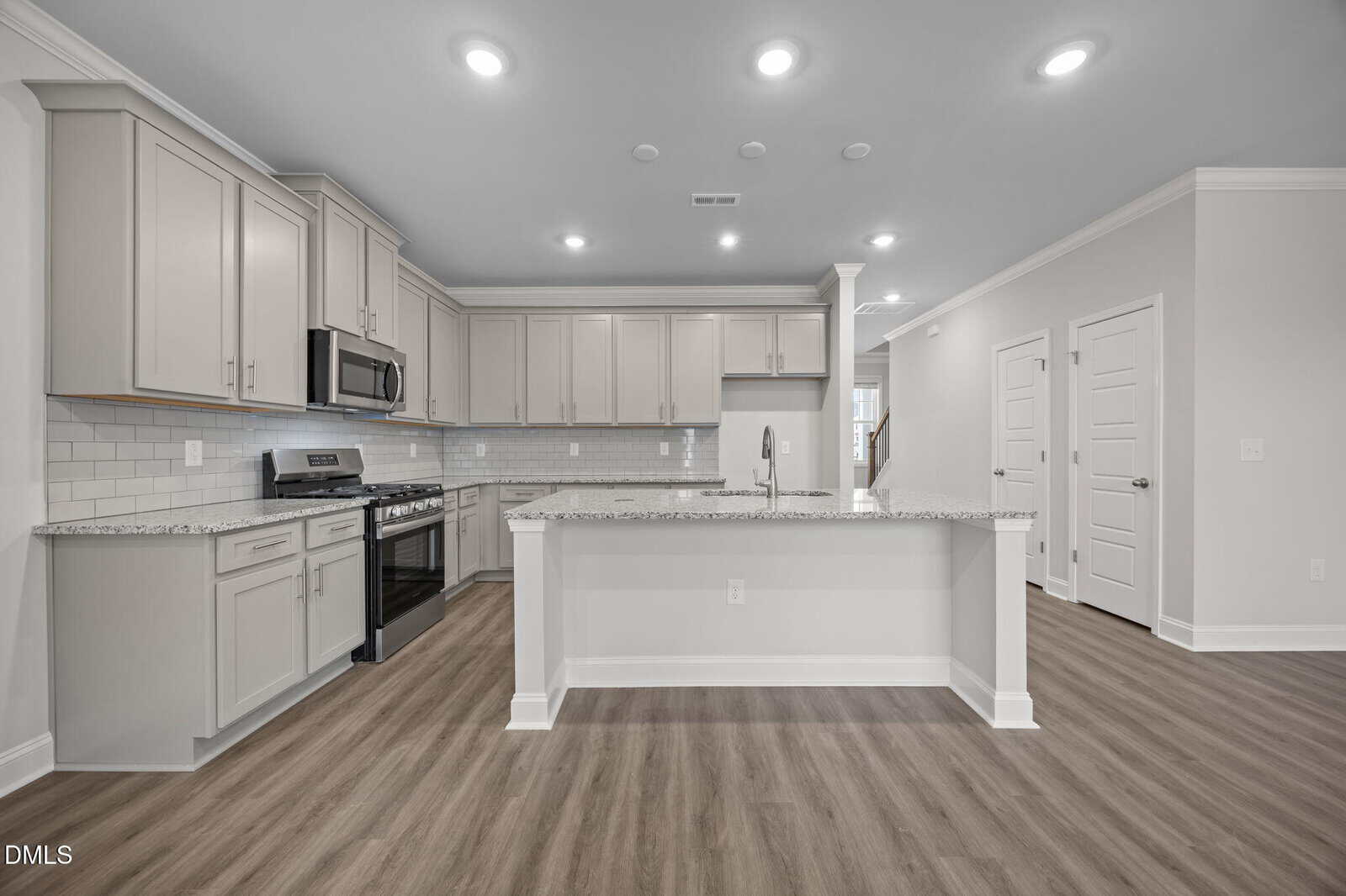Modern kitchen with light gray cabinets, stainless steel appliances, white subway tile backsplash, granite countertops, large island with sink, and wood-look flooring under recessed ceiling lights.