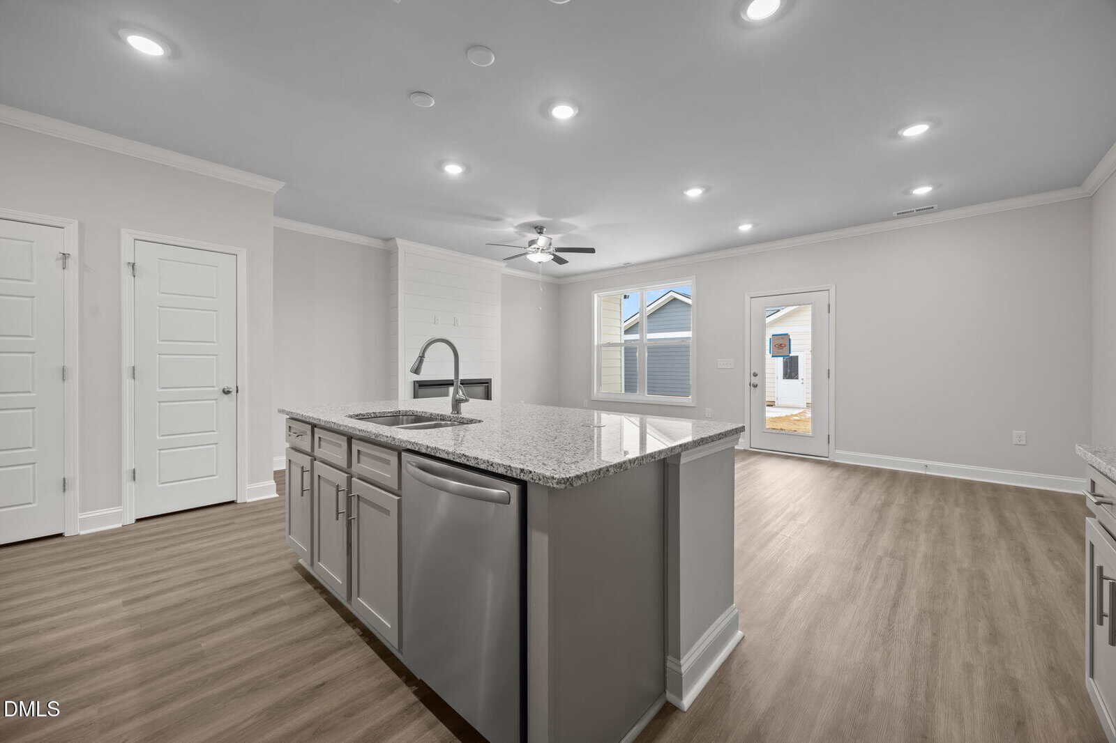 Modern kitchen with light gray cabinets, stainless steel appliances, granite island, wood flooring, and recessed lighting, opening into a bright living area with a ceiling fan and door to the backyard.