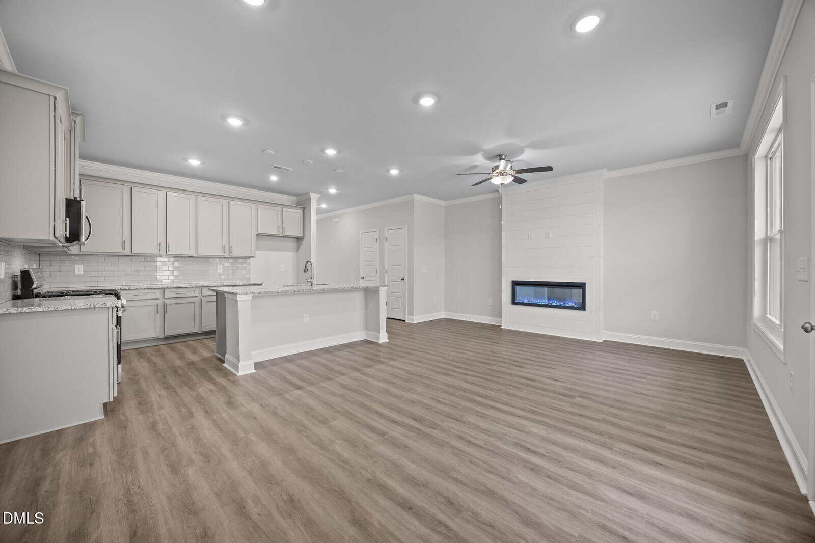 Spacious modern kitchen and living area with light-colored cabinets, granite countertops, wood flooring, built-in electric fireplace, recessed lighting, and large windows letting in natural light.
