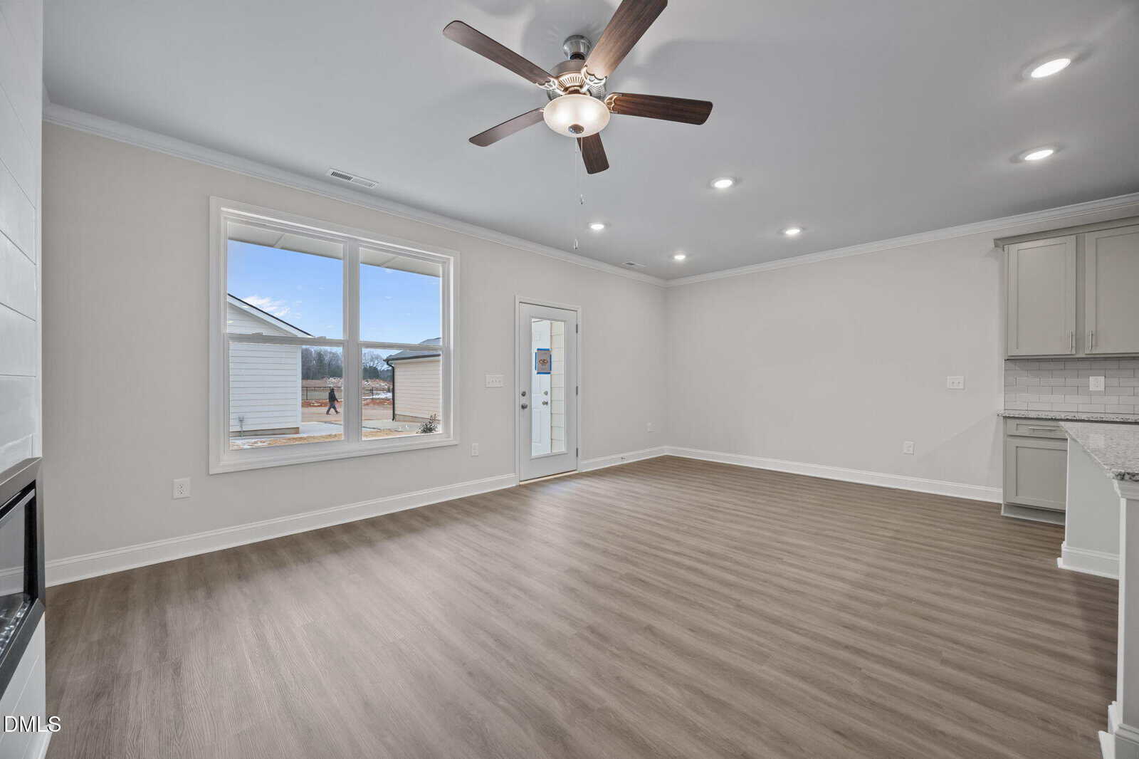 Spacious, empty living room with light wood flooring, large window, ceiling fan with lights, recessed ceiling lights, and a door leading outside. A portion of a kitchen with light cabinets is visible on the right.