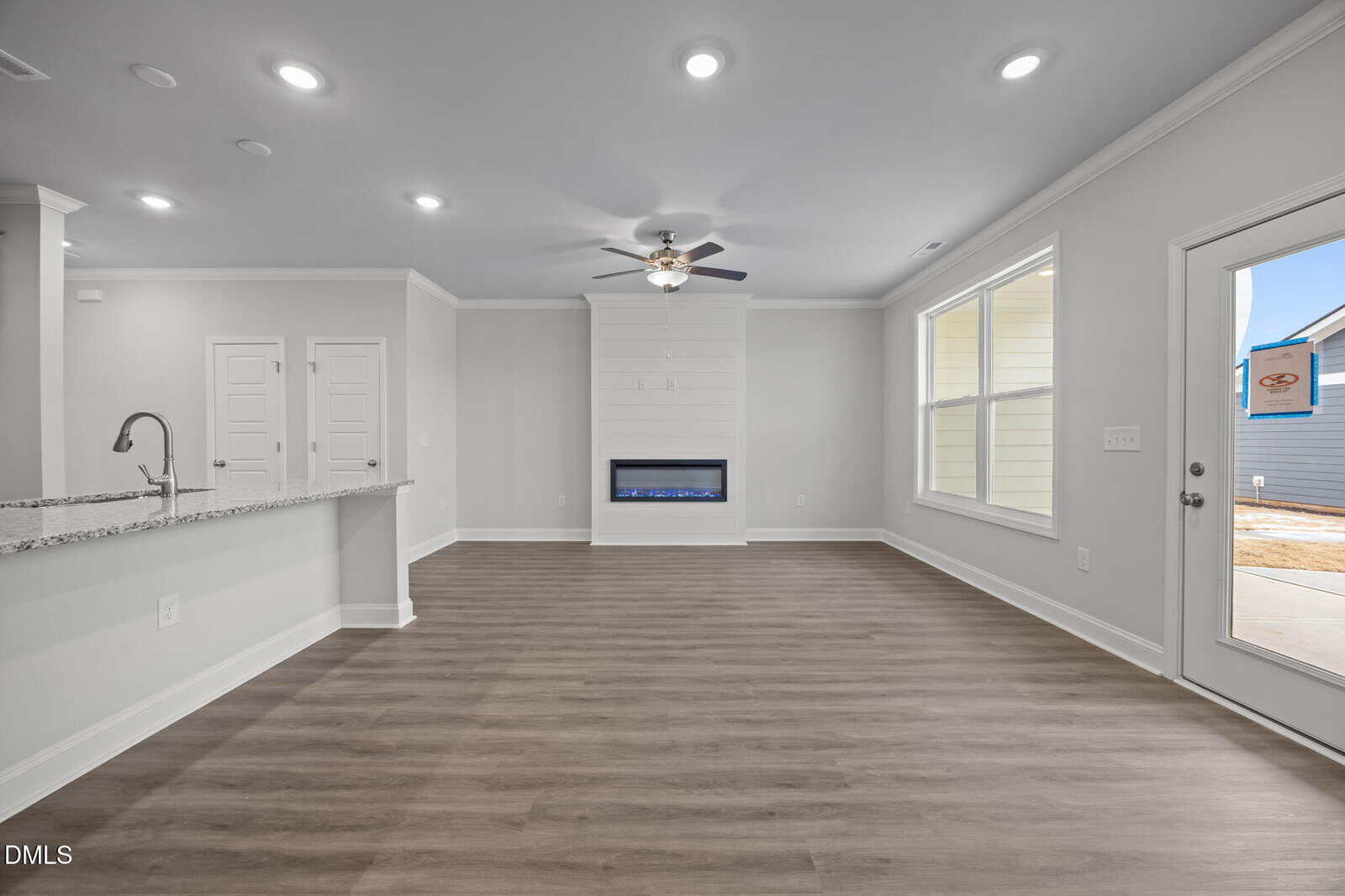A modern, unfurnished living room with light gray walls, wood flooring, recessed lighting, a ceiling fan, a built-in electric fireplace, and large windows letting in natural light. A kitchen counter with a sink is visible on the left.